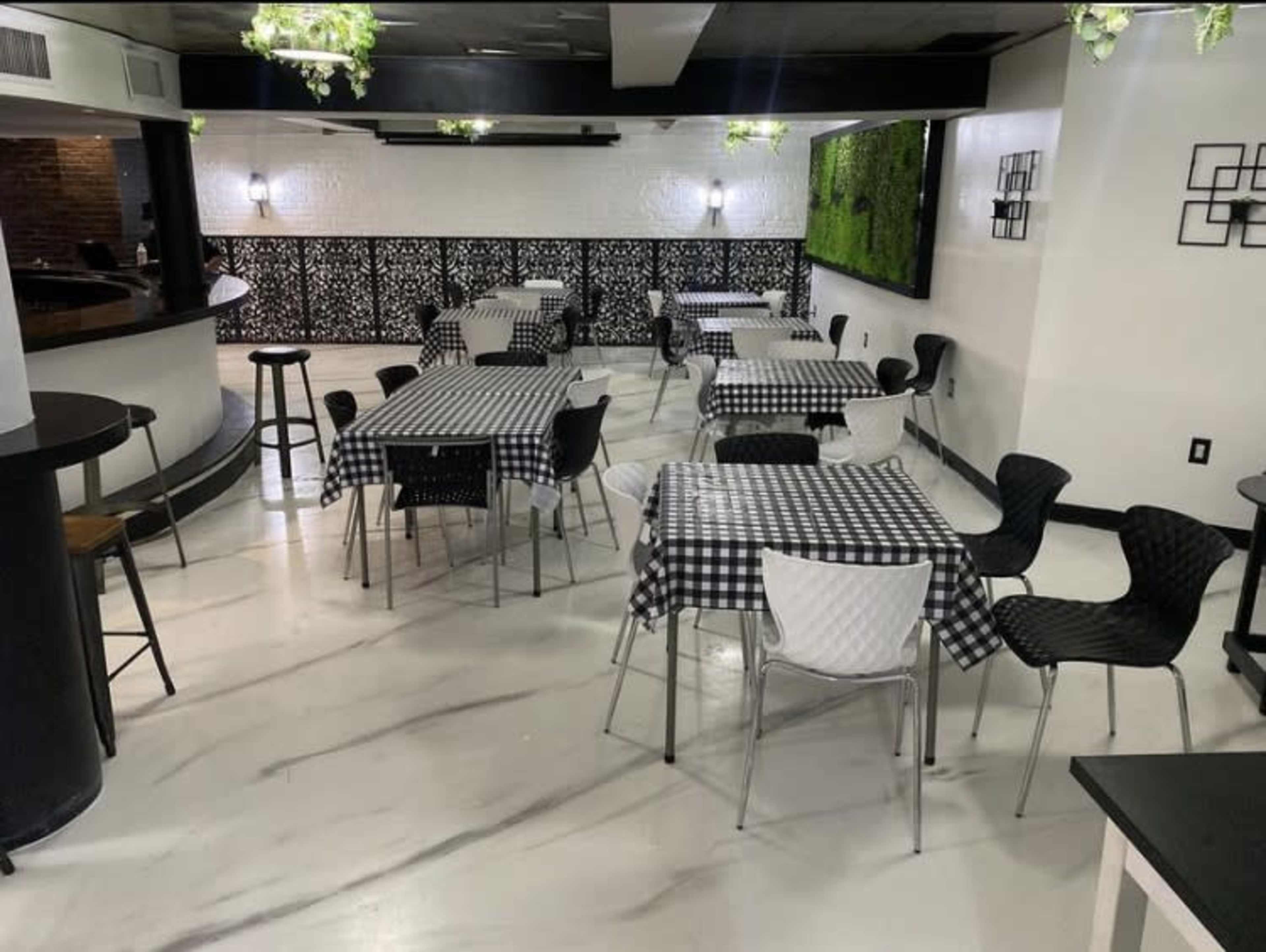 A restaurant interior with several tables covered in black and white checked tablecloths, surrounded by modern seating and greenery decor on the walls.