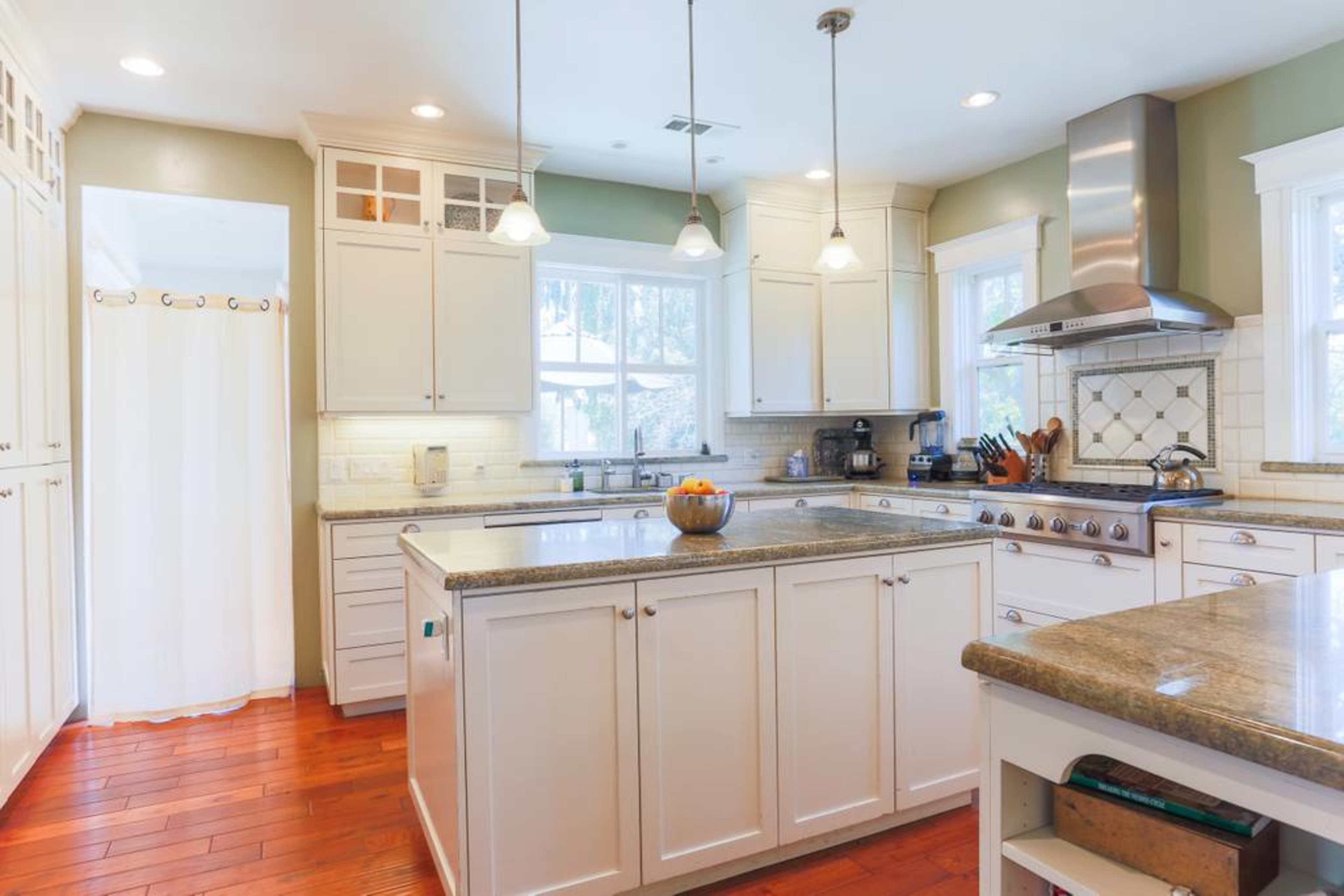 A bright kitchen features white cabinetry, a gray stone island with a bowl of fruit, and wooden flooring.