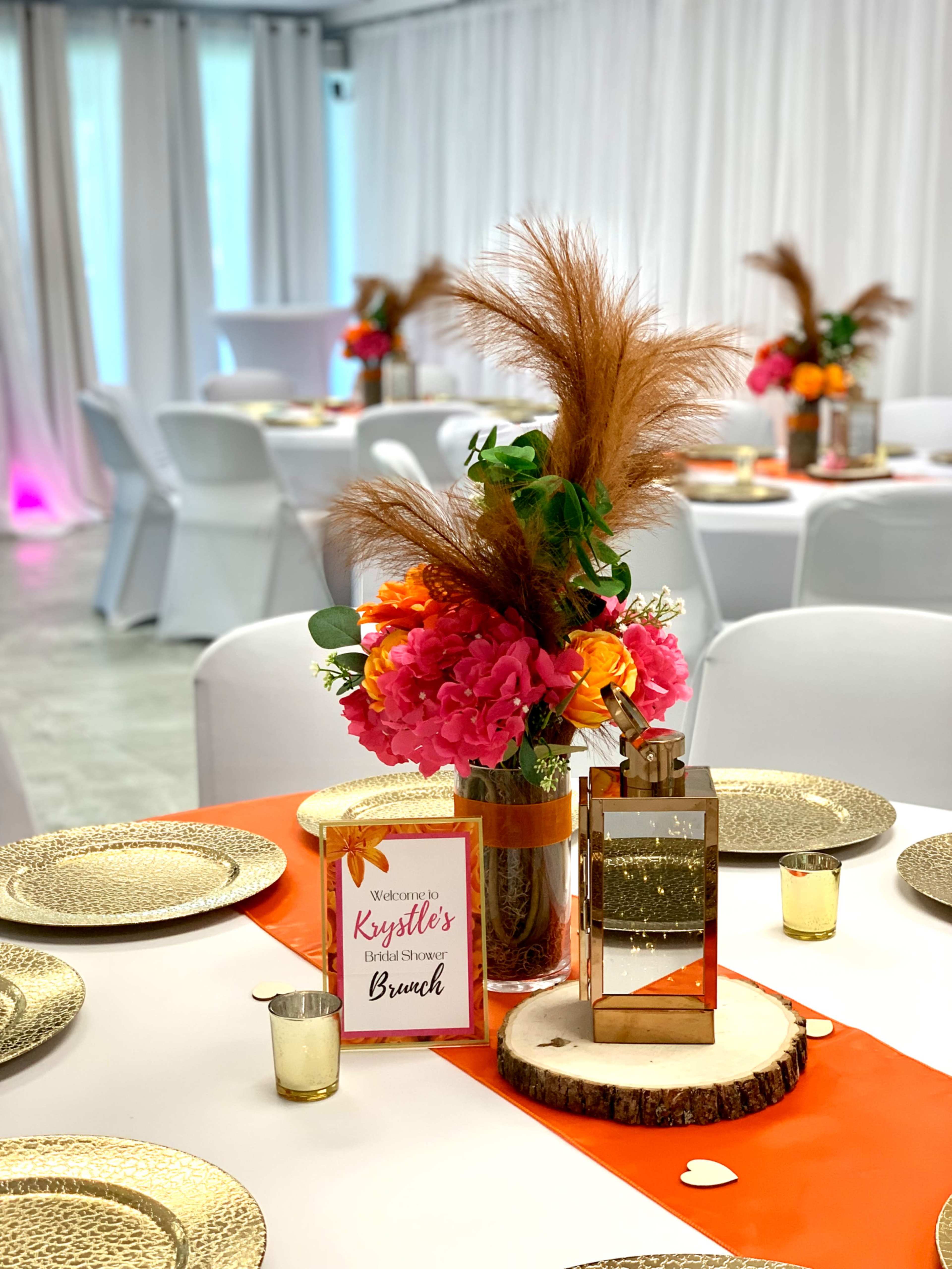 A decorated dining table features a floral centerpiece with pink and orange flowers, a mirrored lantern, and a welcome sign for a brunch event.