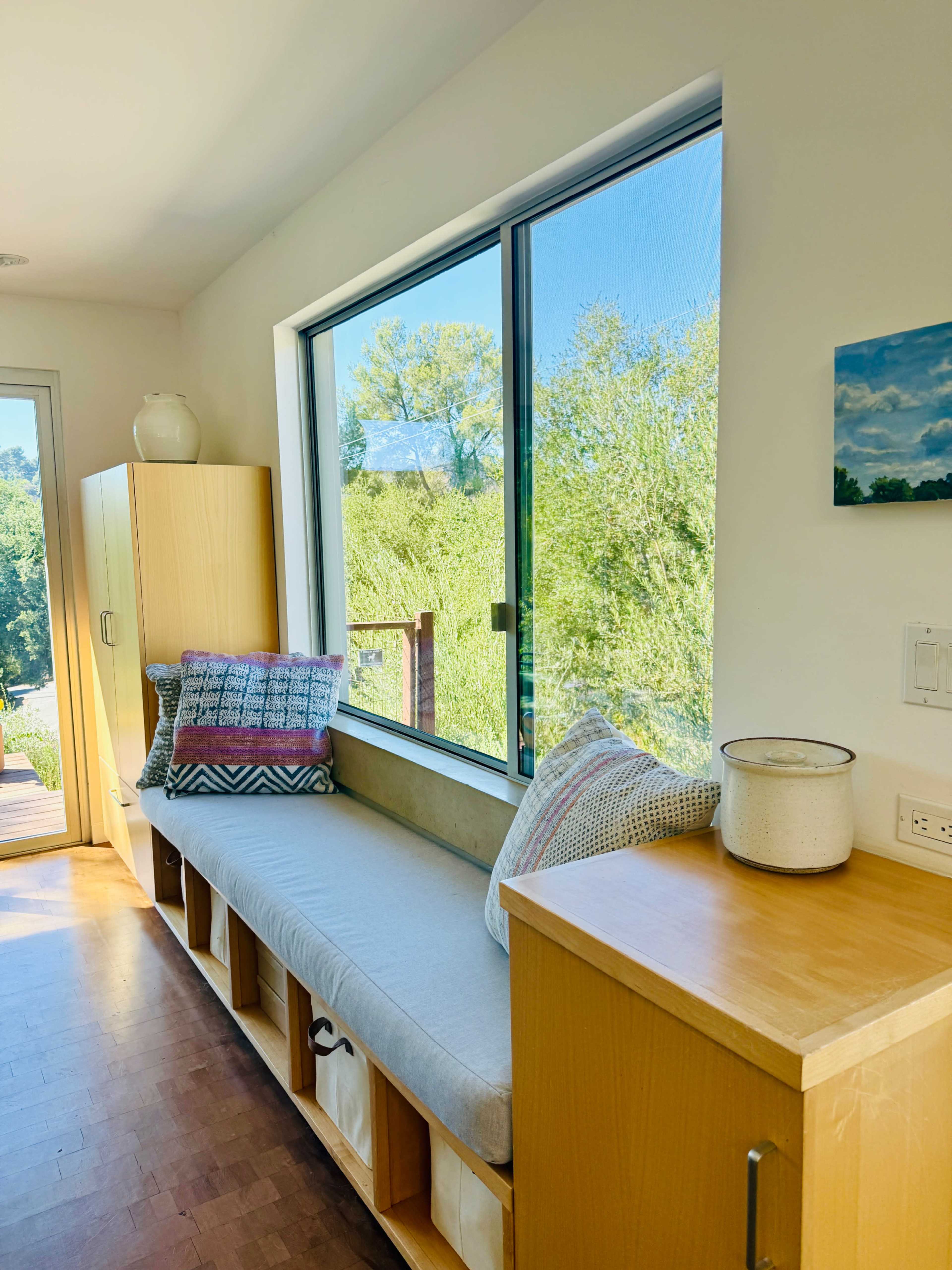 A bright nook with a cushioned bench sits beside a large window, showcasing greenery outside.