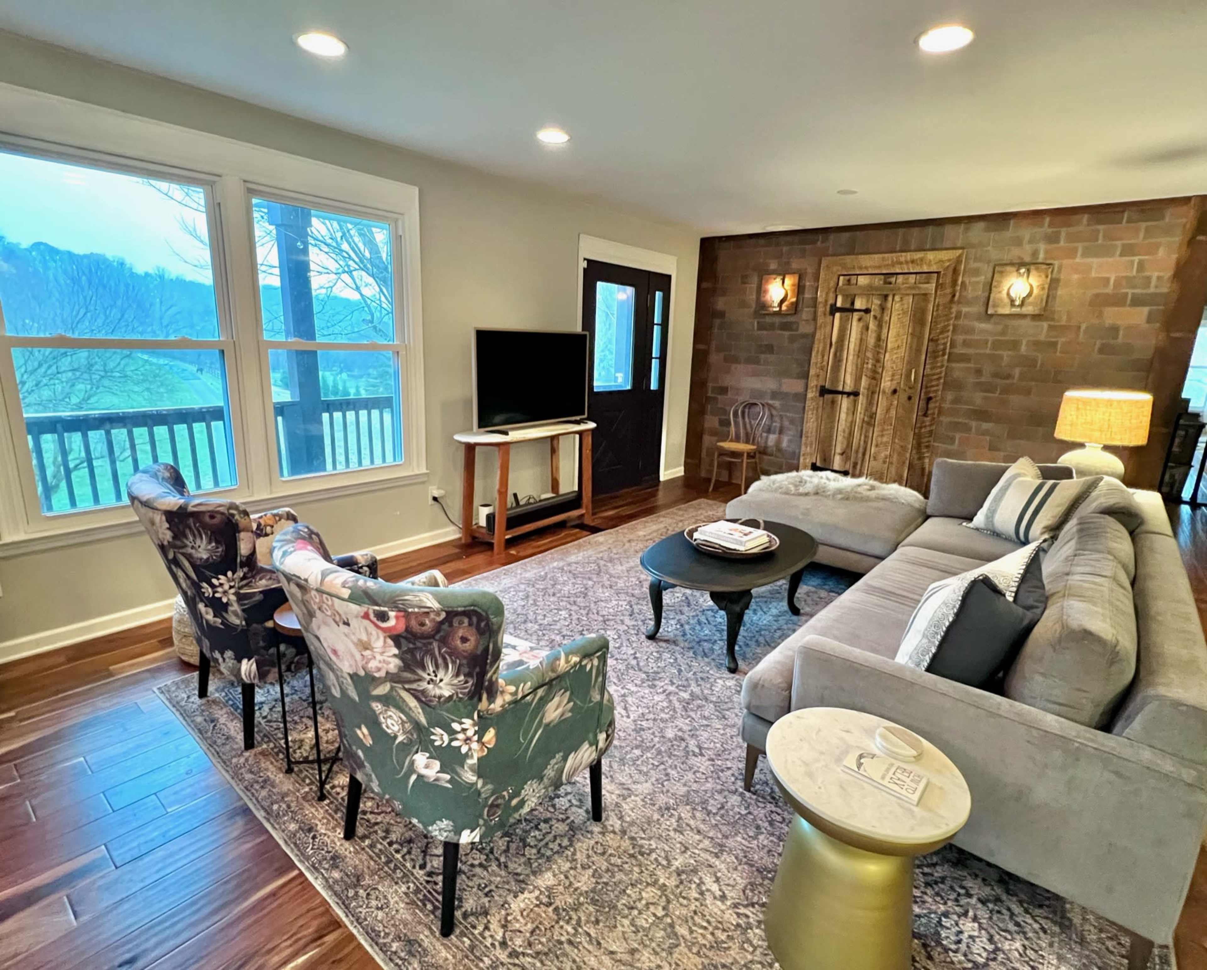 A cozy living room with a sectional sofa, two floral armchairs, a coffee table, a television, and large windows overlooking a scenic view.
