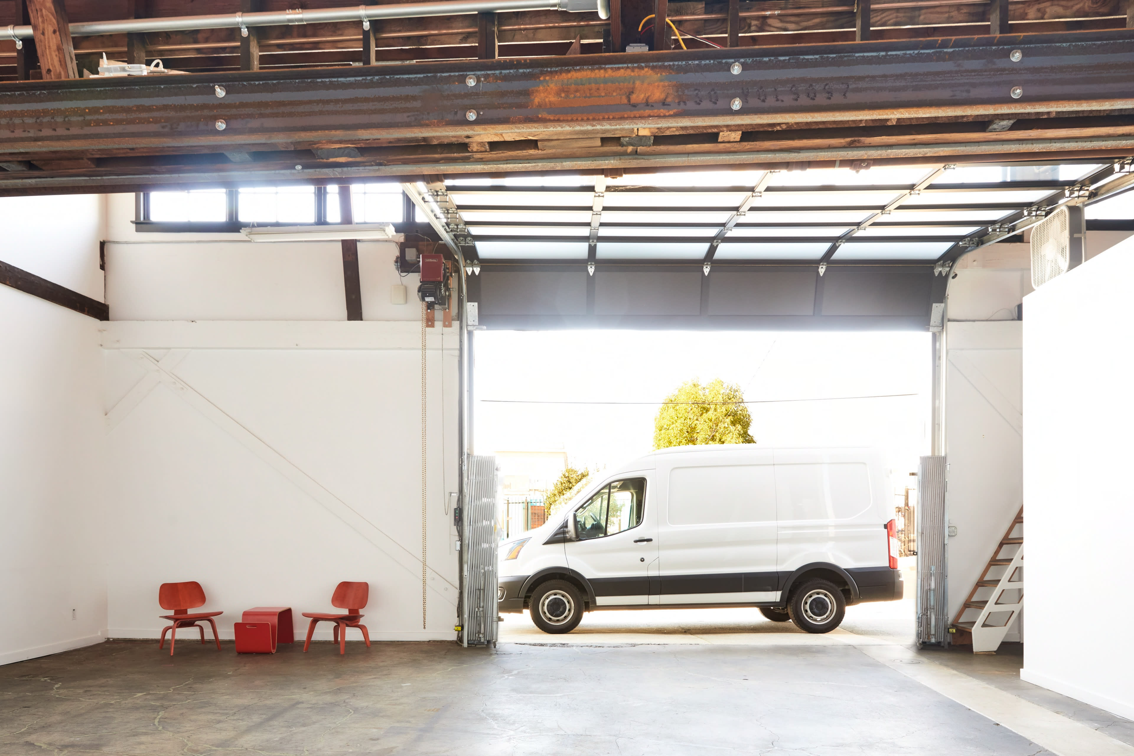 A white delivery van is parked outside an open garage door, with two red chairs positioned inside the empty space.