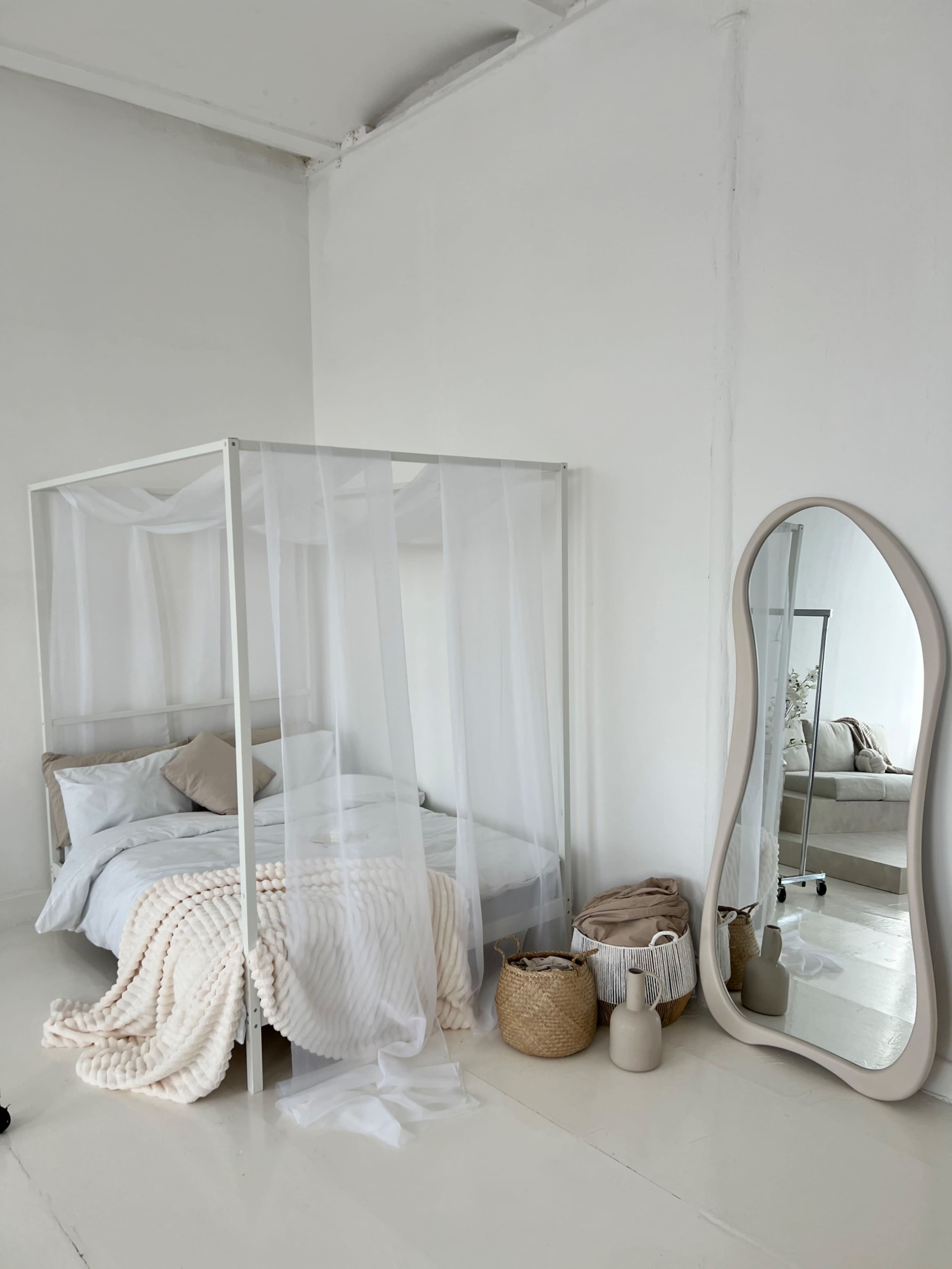 The image shows a minimalist bedroom featuring a four-poster bed with sheer white curtains, a large mirror, and woven baskets placed nearby.