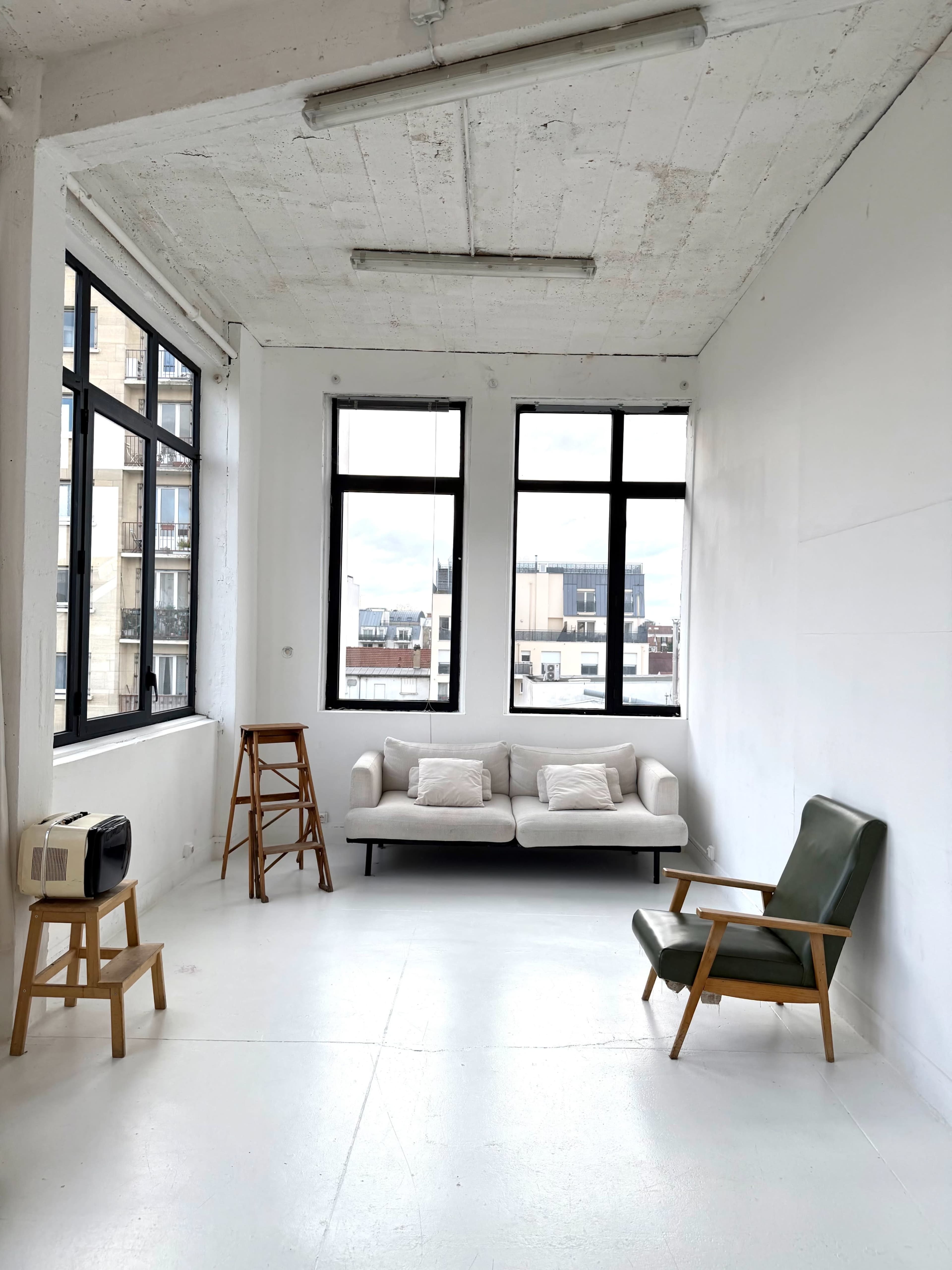 The image shows a sparsely furnished room with large windows, a light-colored sofa, a green chair, and a small vintage radio.