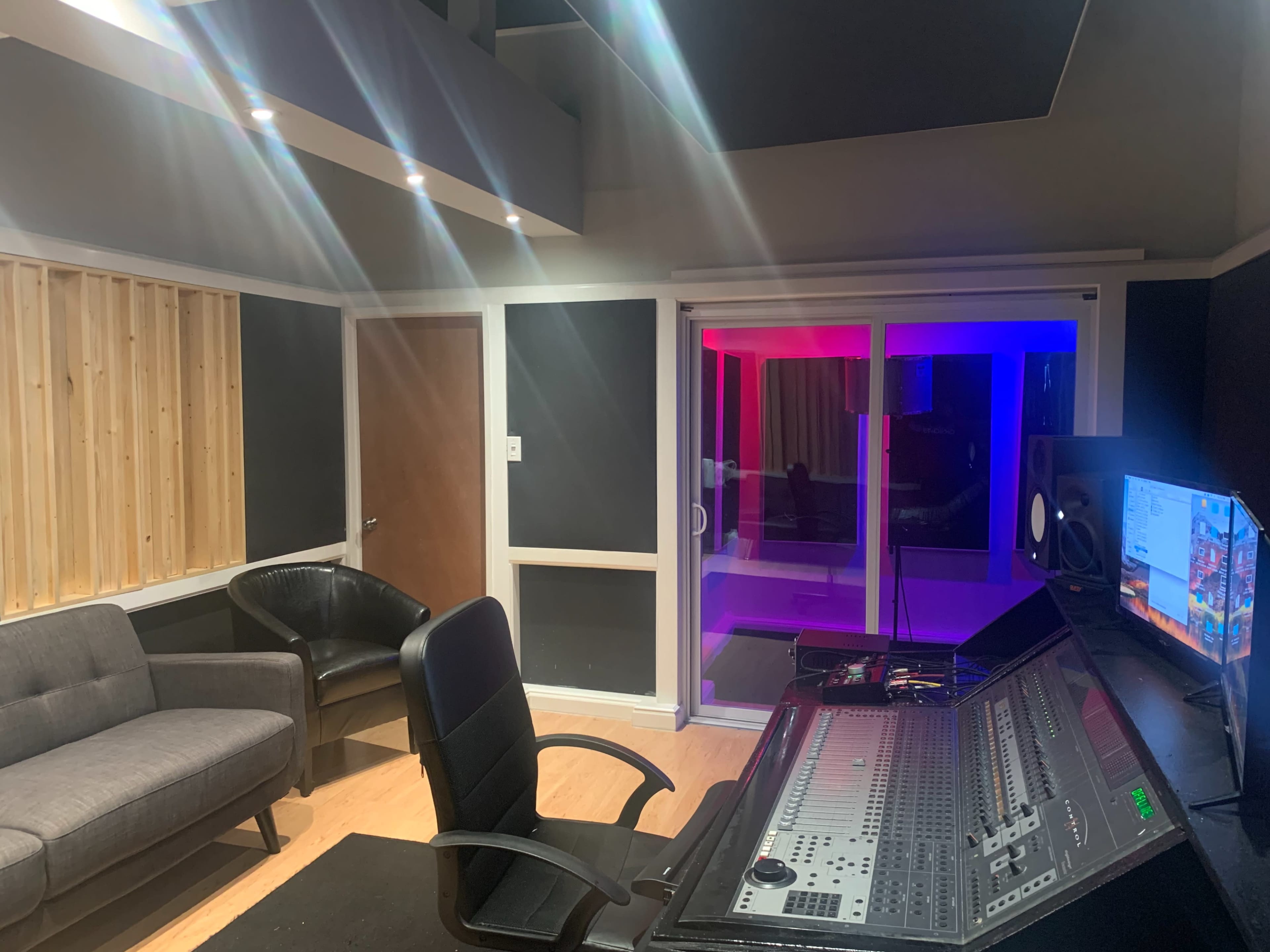 A music studio with a mixing console, a black office chair, a gray couch, and a glass-paneled room illuminated with colored lights.