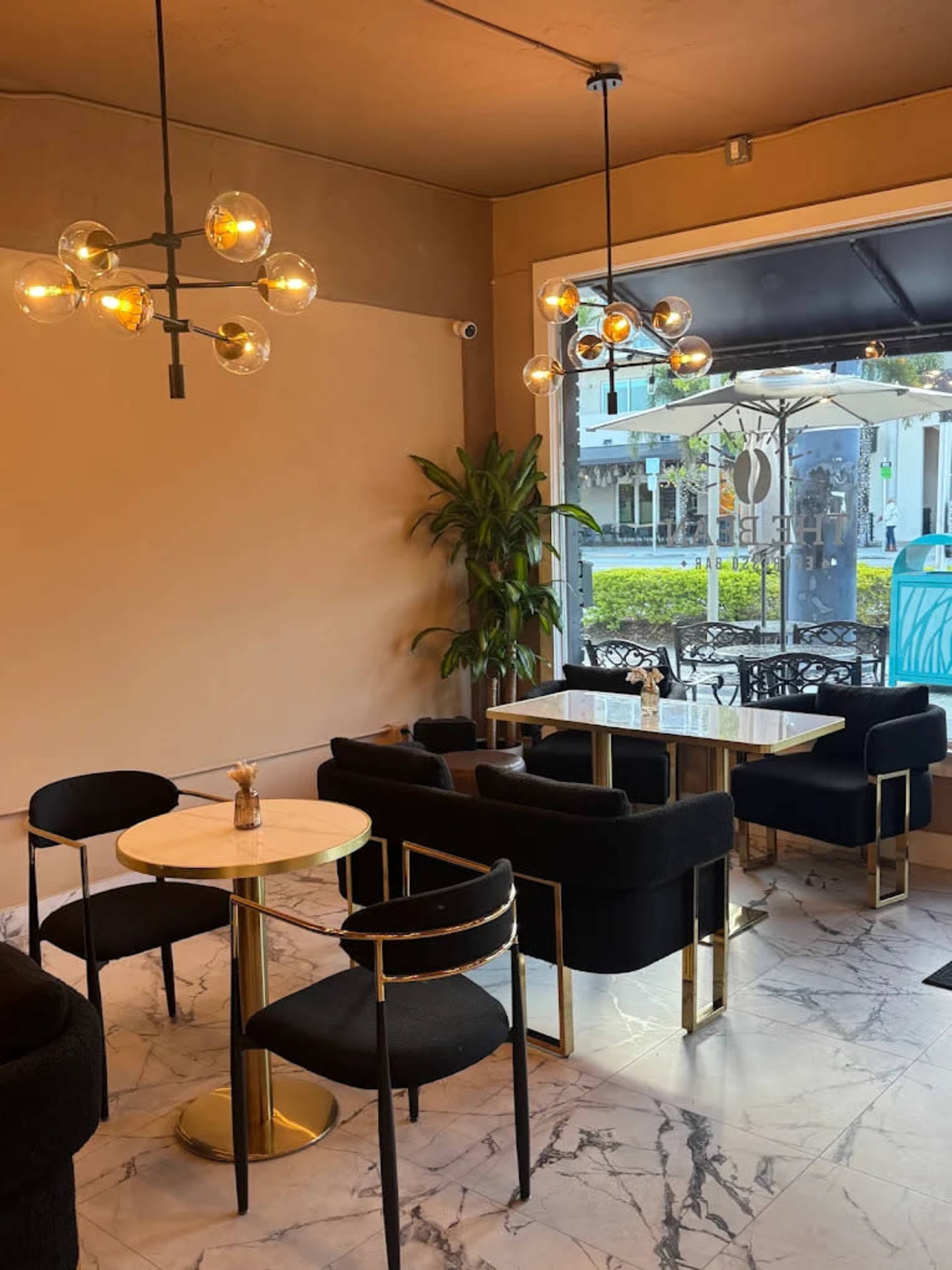 The image shows a modern café with black seating, a round table, and a large window overlooking an outdoor area with greenery.