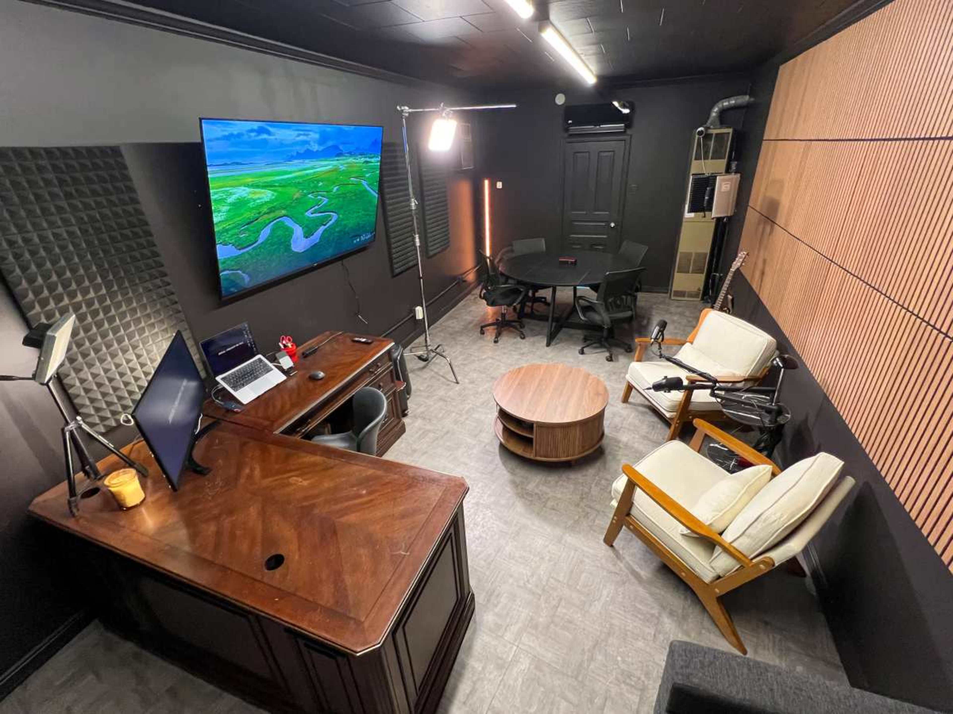 The image shows an office space featuring a wooden desk, a large wall-mounted screen displaying a landscape, a round coffee table, two armchairs, and a dark conference table illuminated by a standing light.