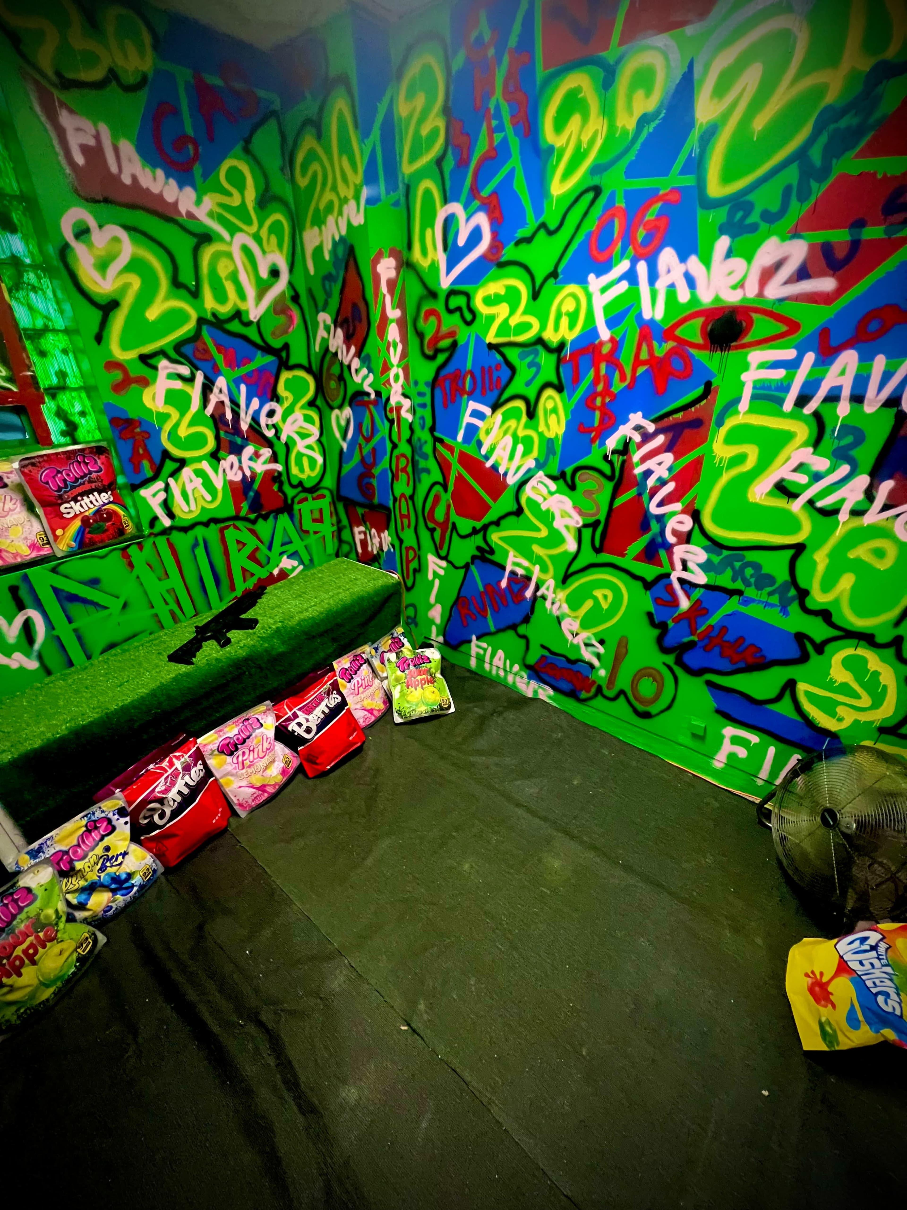 The image depicts a small room with vibrant green walls covered in colorful graffiti and a light green bench, surrounded by bags of snacks on the floor.