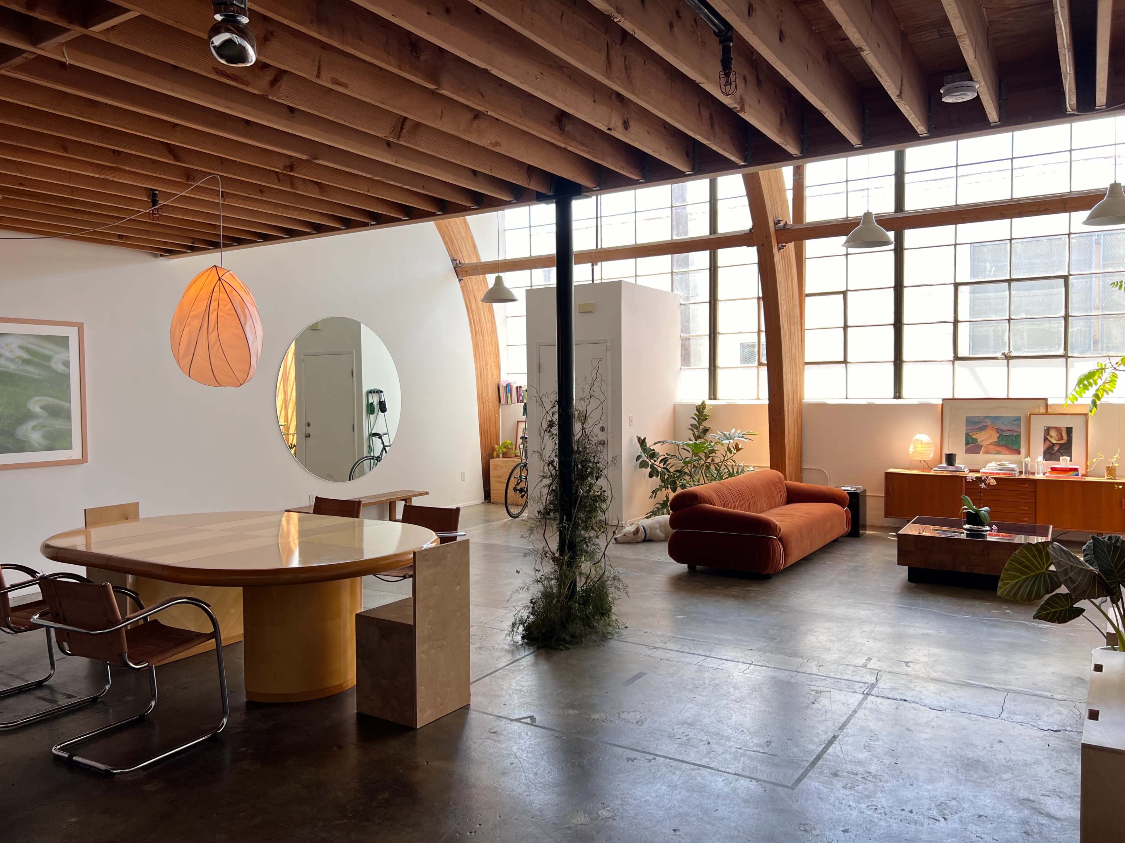 The room features a modern open layout with a large round table, a sofa, and various plants, all situated under exposed wooden beams and large windows.