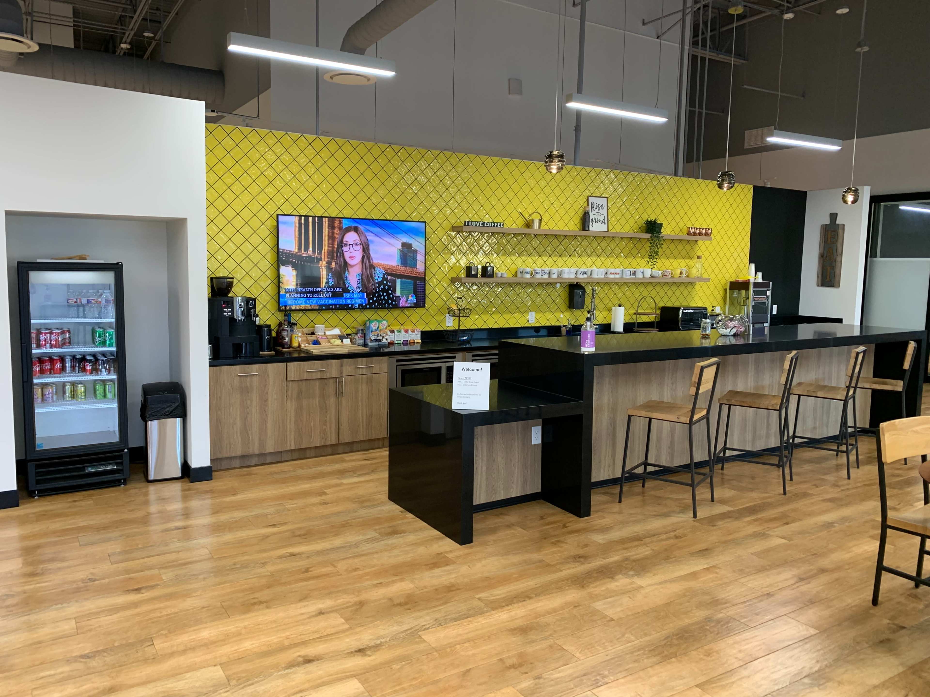 A modern kitchen space featuring a long black countertop, wooden shelves with various items, a television mounted on a yellow wall, and a refrigerator filled with drinks.
