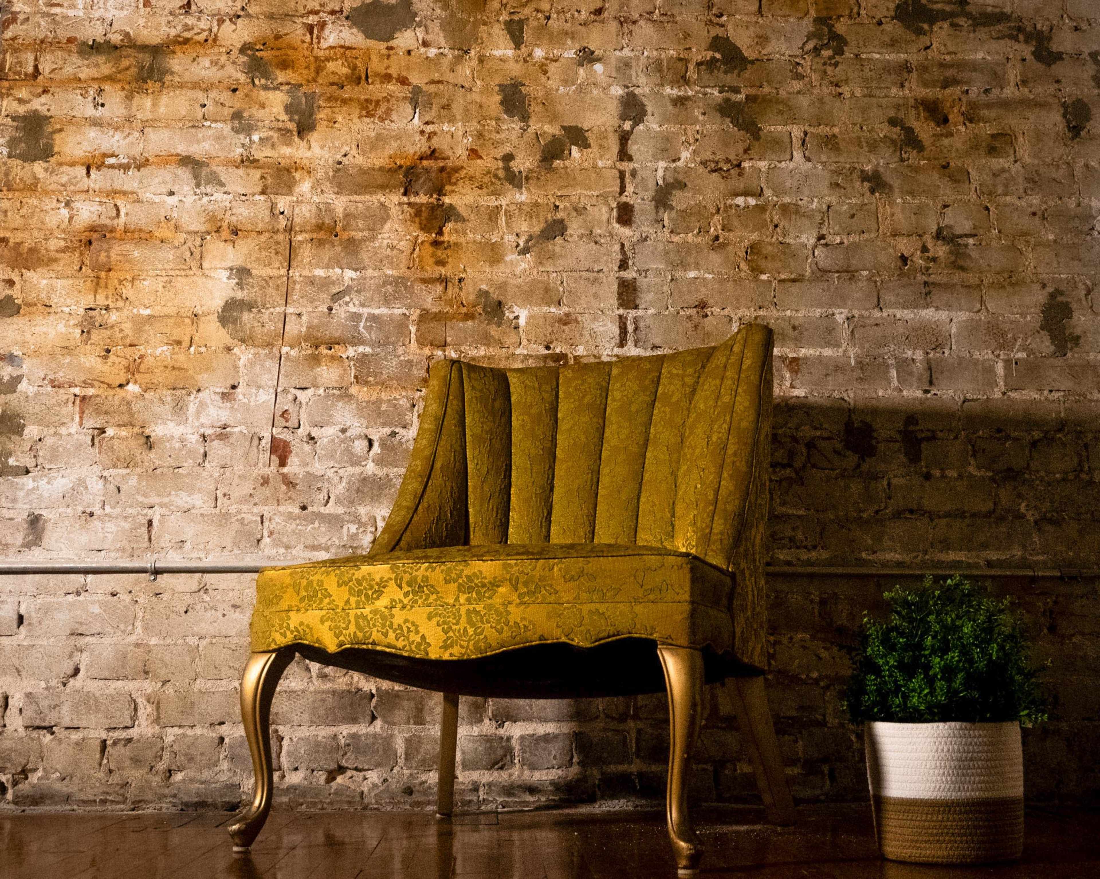 A yellow upholstered chair with a curved backrest sits against a textured brick wall, next to a potted plant.