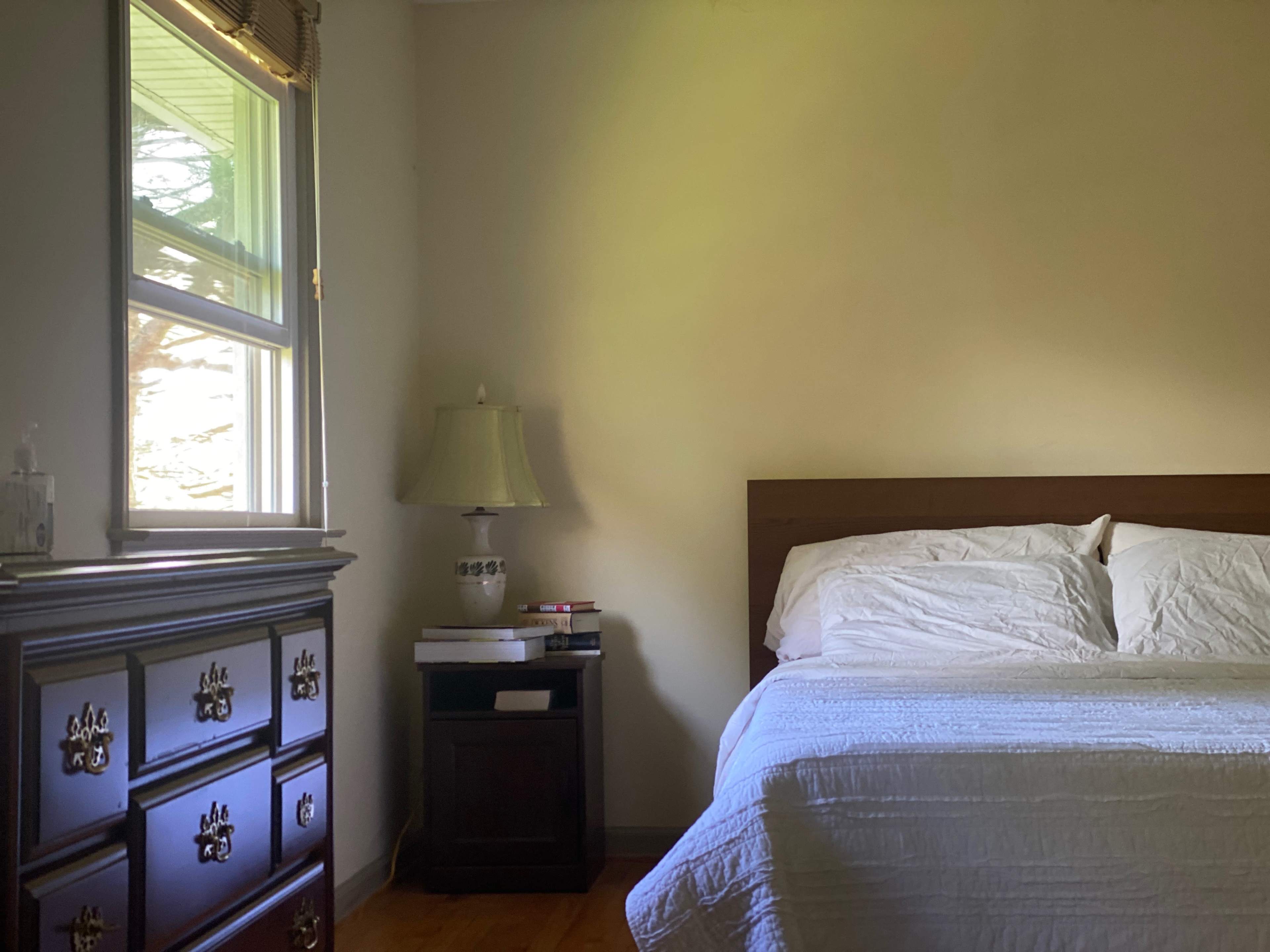 The image shows a bedroom with a bed, a wooden dresser, a lamp on a nightstand, and a window allowing natural light.