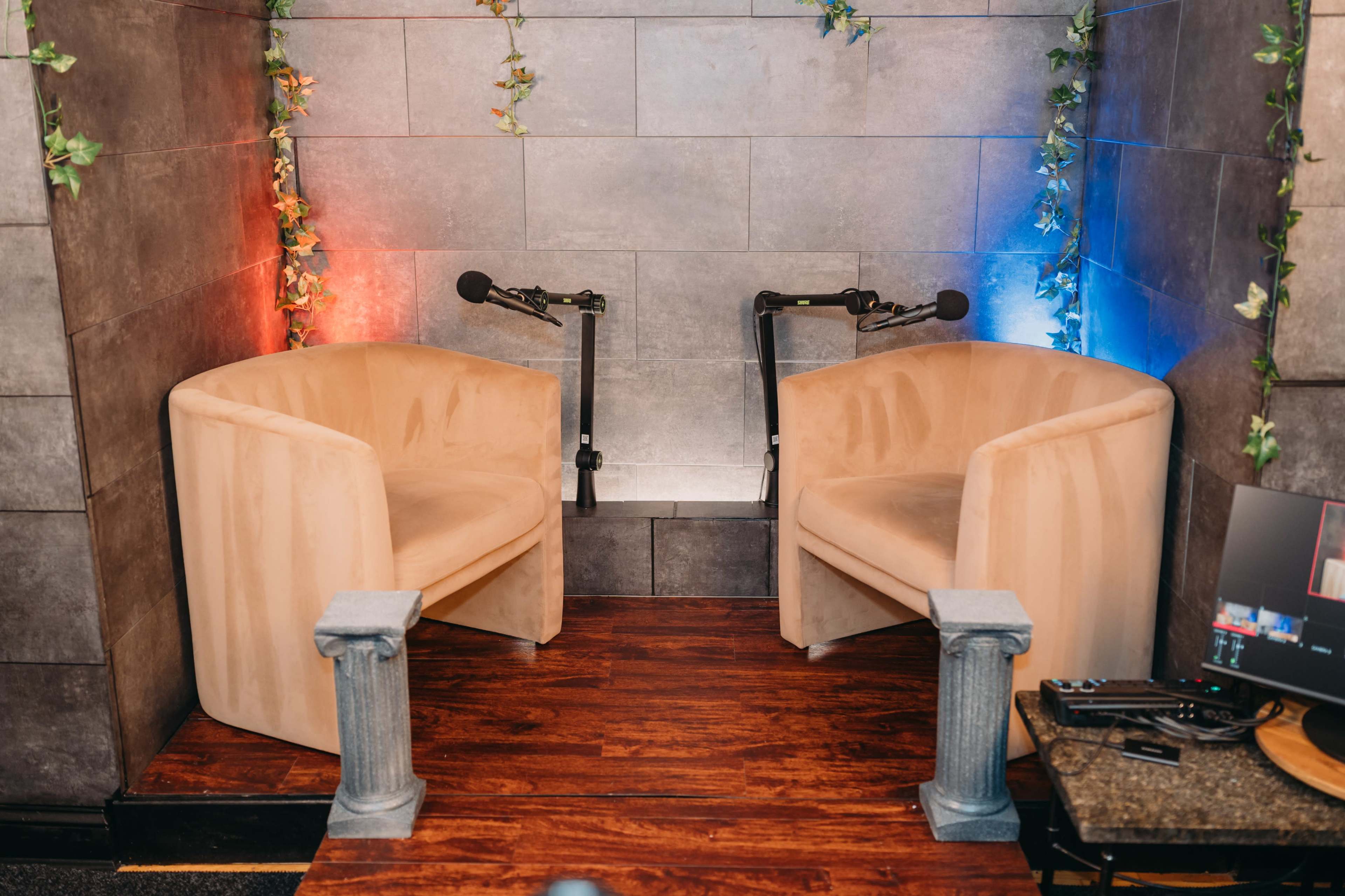 The image shows a small podcast or interview setup featuring two beige chairs with microphones positioned in front of them, adorned with greenery on the walls.
