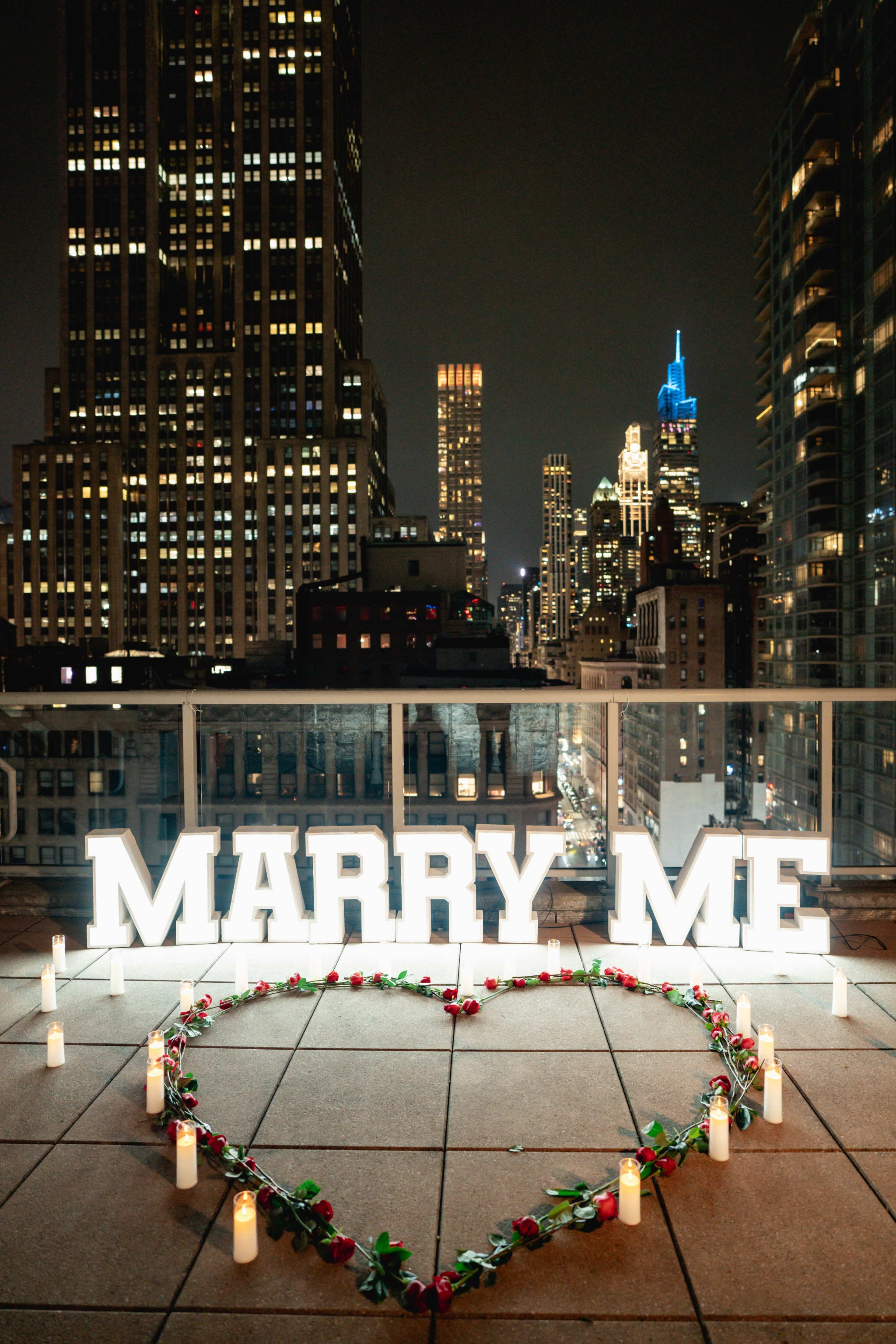 The image shows a rooftop proposal setup at night, featuring illuminated letters spelling "MARRY ME" and a heart-shaped arrangement of candles and rose petals with a city skyline in the background.