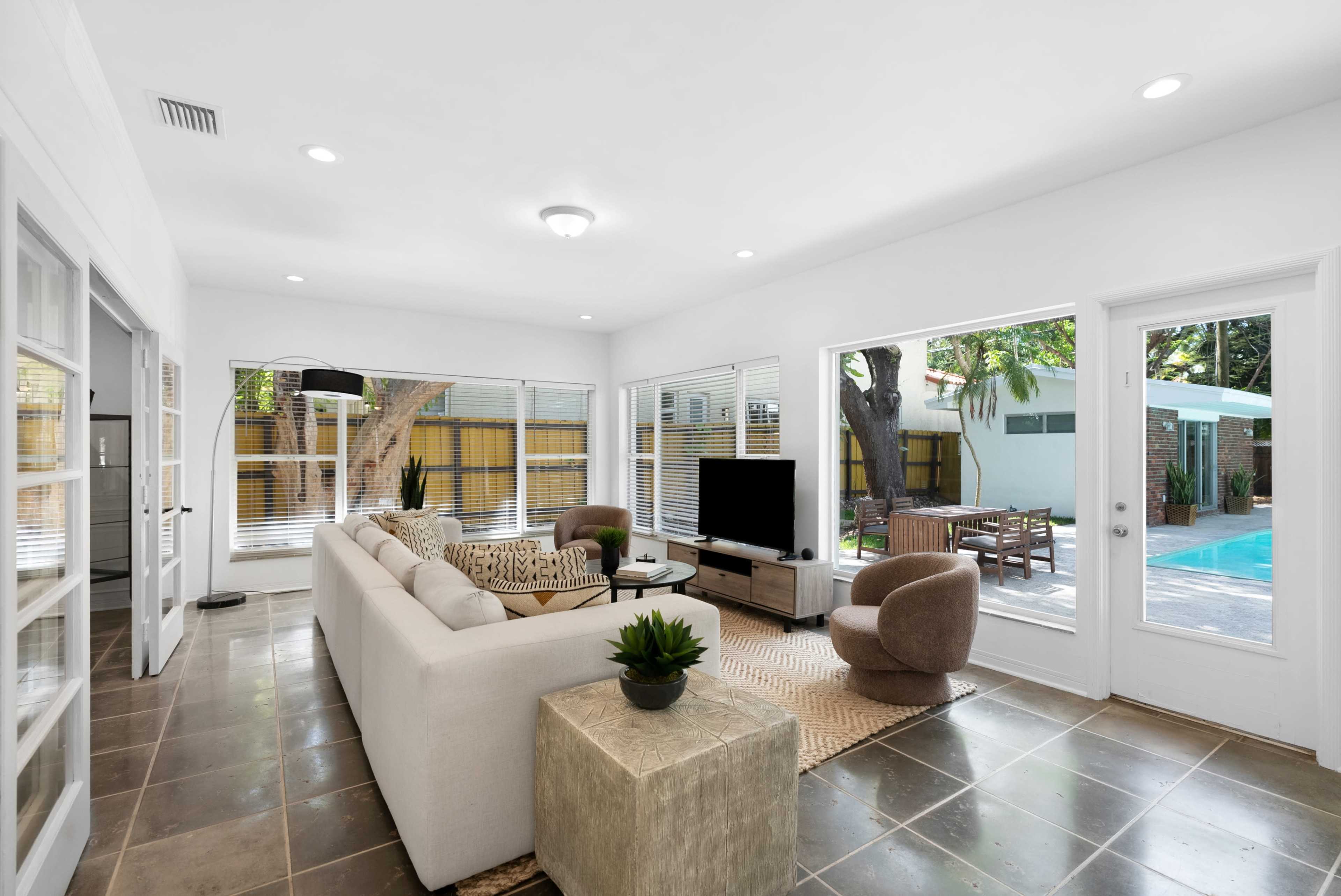 The image shows a modern living room with a large white sofa, a television, and windows that open to a patio and pool area.