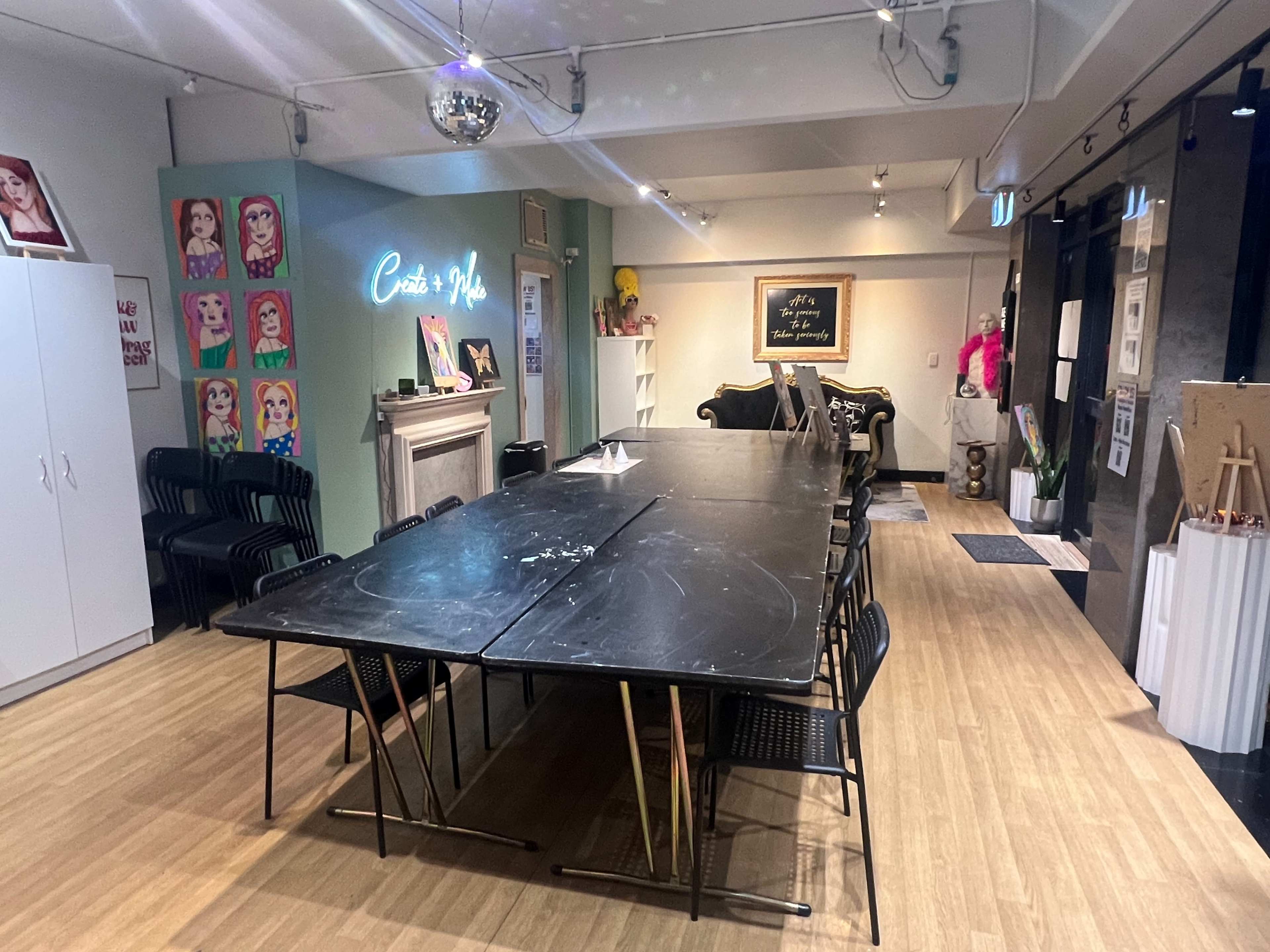 The image shows a spacious room featuring a long black table surrounded by black chairs, with colorful artwork on the walls and a disco ball hanging from the ceiling.