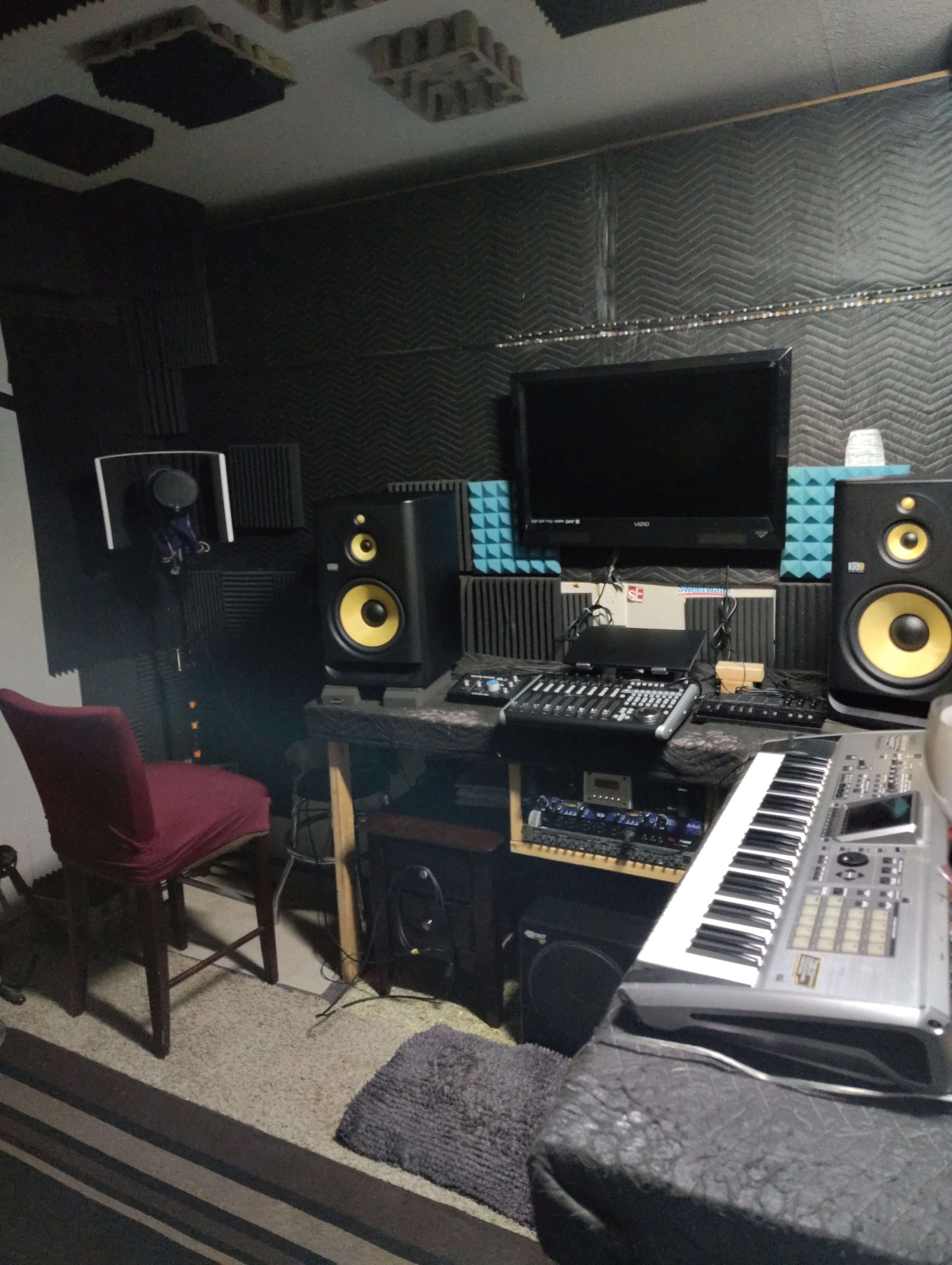 The image shows a recording studio with a microphone on a stand, two large studio monitors, a mixer, and a keyboard positioned on a desk.