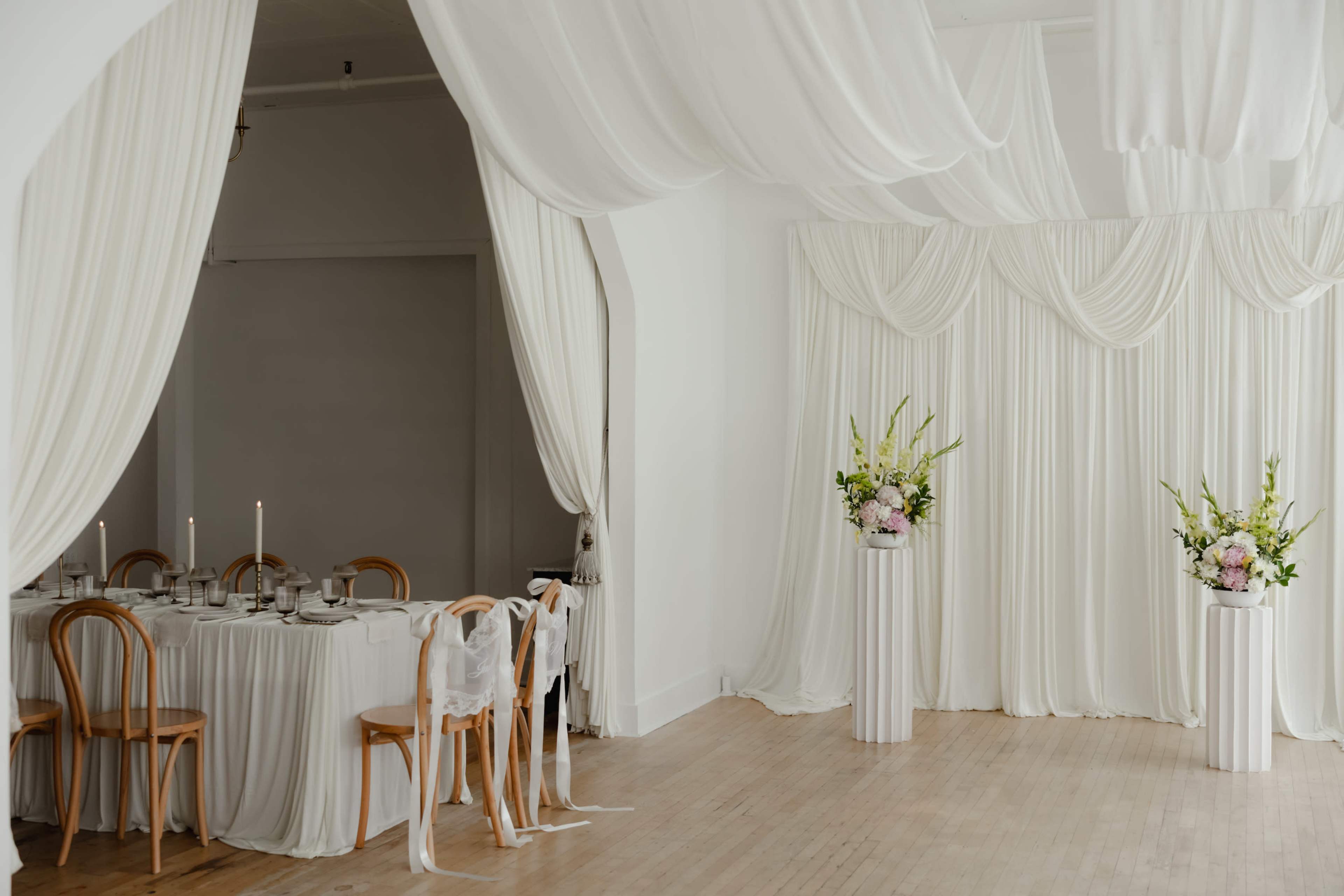 The image shows a spacious, elegantly decorated room with white drapery, a dining setup with candles, and floral arrangements on pedestals.