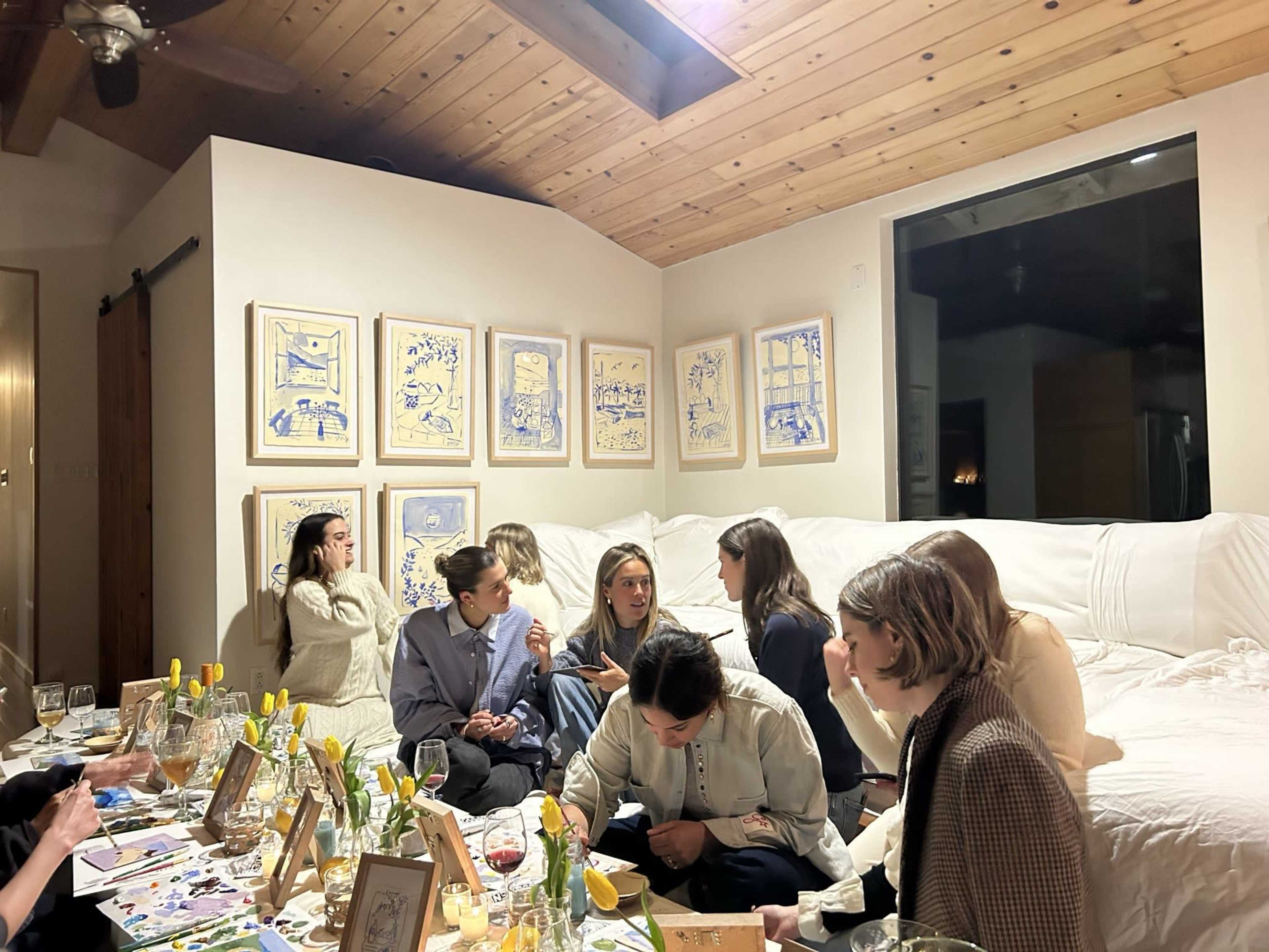 A group of women is gathered in a warmly lit room, seated on a white sofa and engaging in conversation, with artwork displayed on the walls.