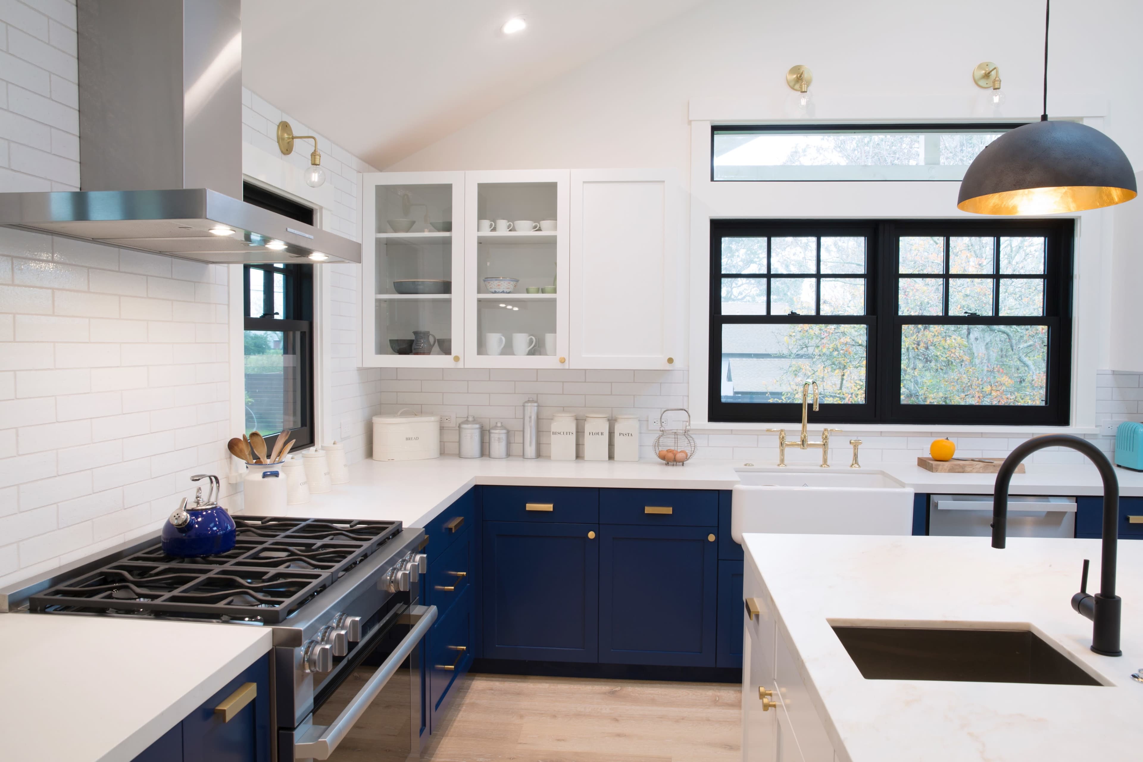 A modern kitchen features blue and white cabinetry, a stainless steel gas stove, and large windows providing natural light.