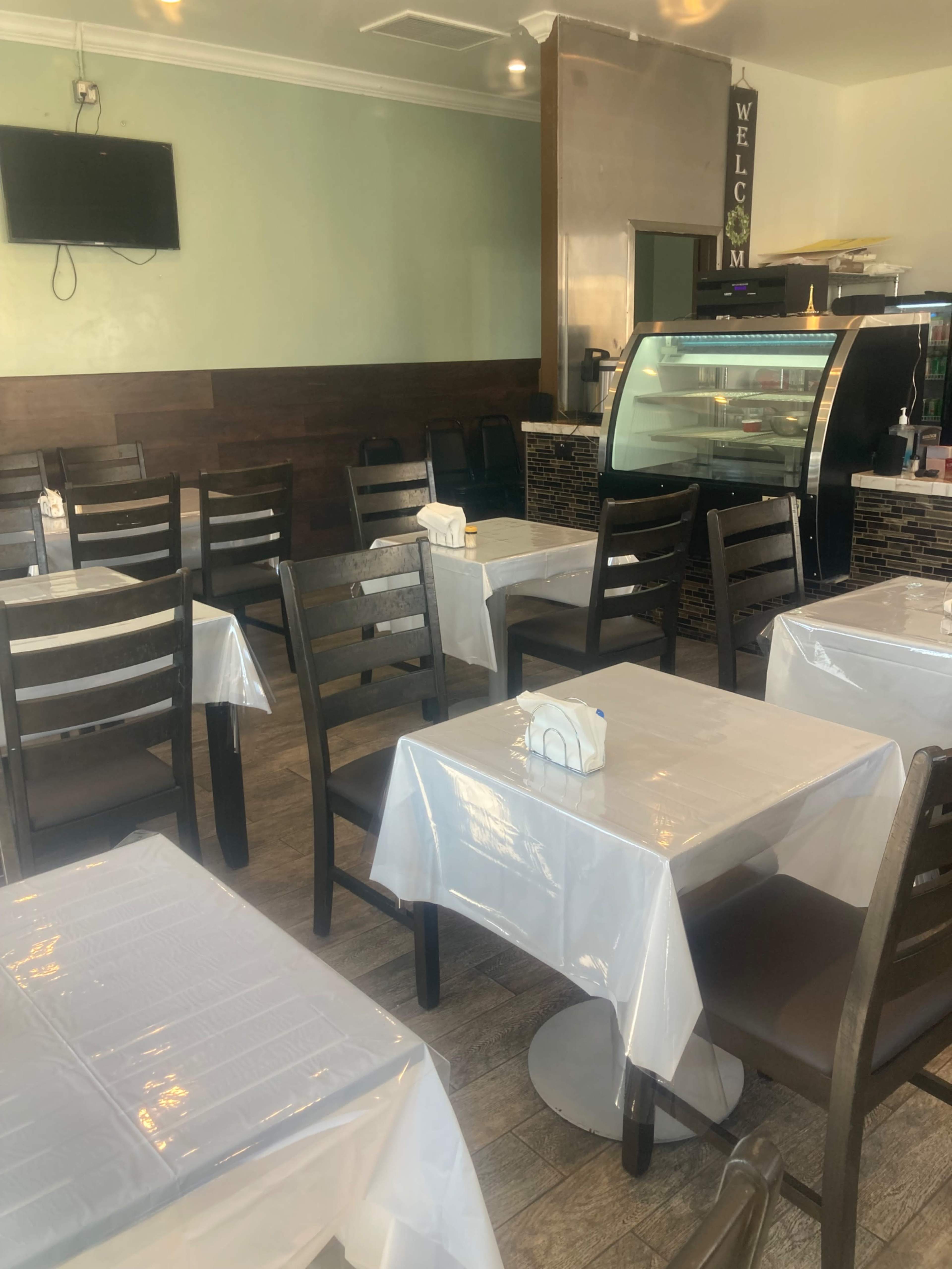The image shows the interior of a restaurant featuring wooden tables and chairs, with white tablecloths and a counter displaying a glass case.