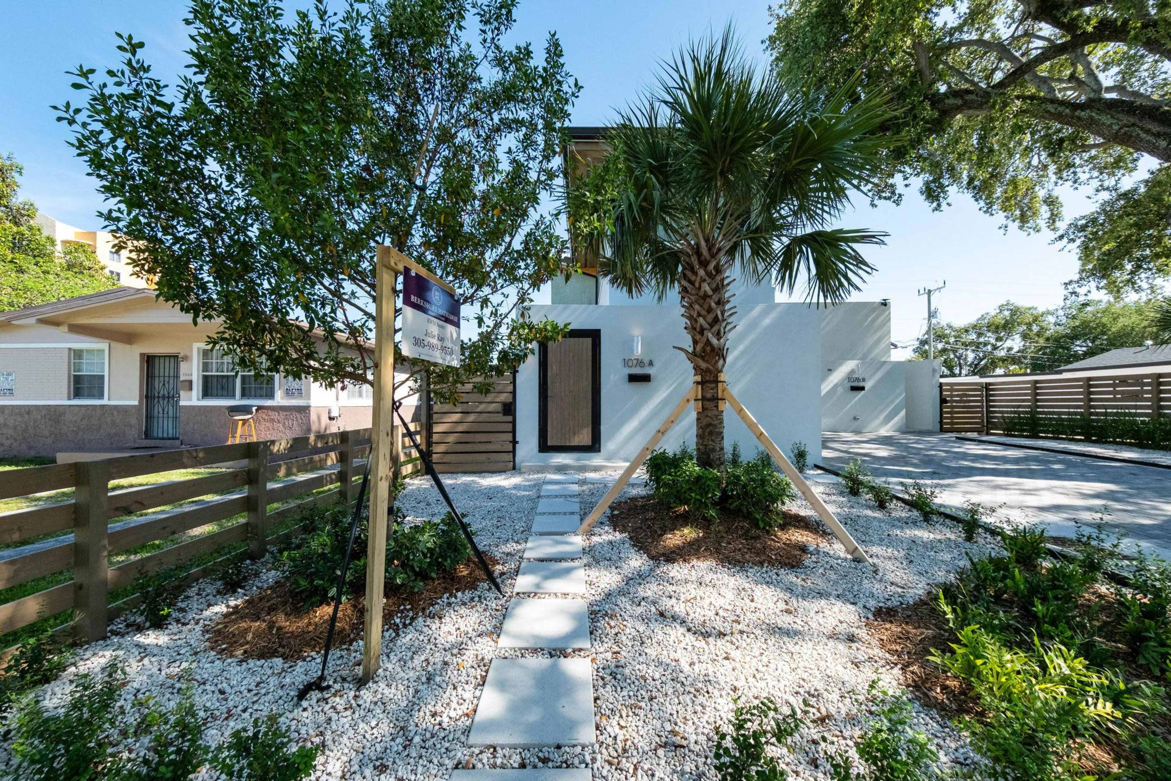 The image shows a modern white house with a palm tree and landscaped gravel area in the front yard, alongside a wooden fence and a "For Sale" sign.