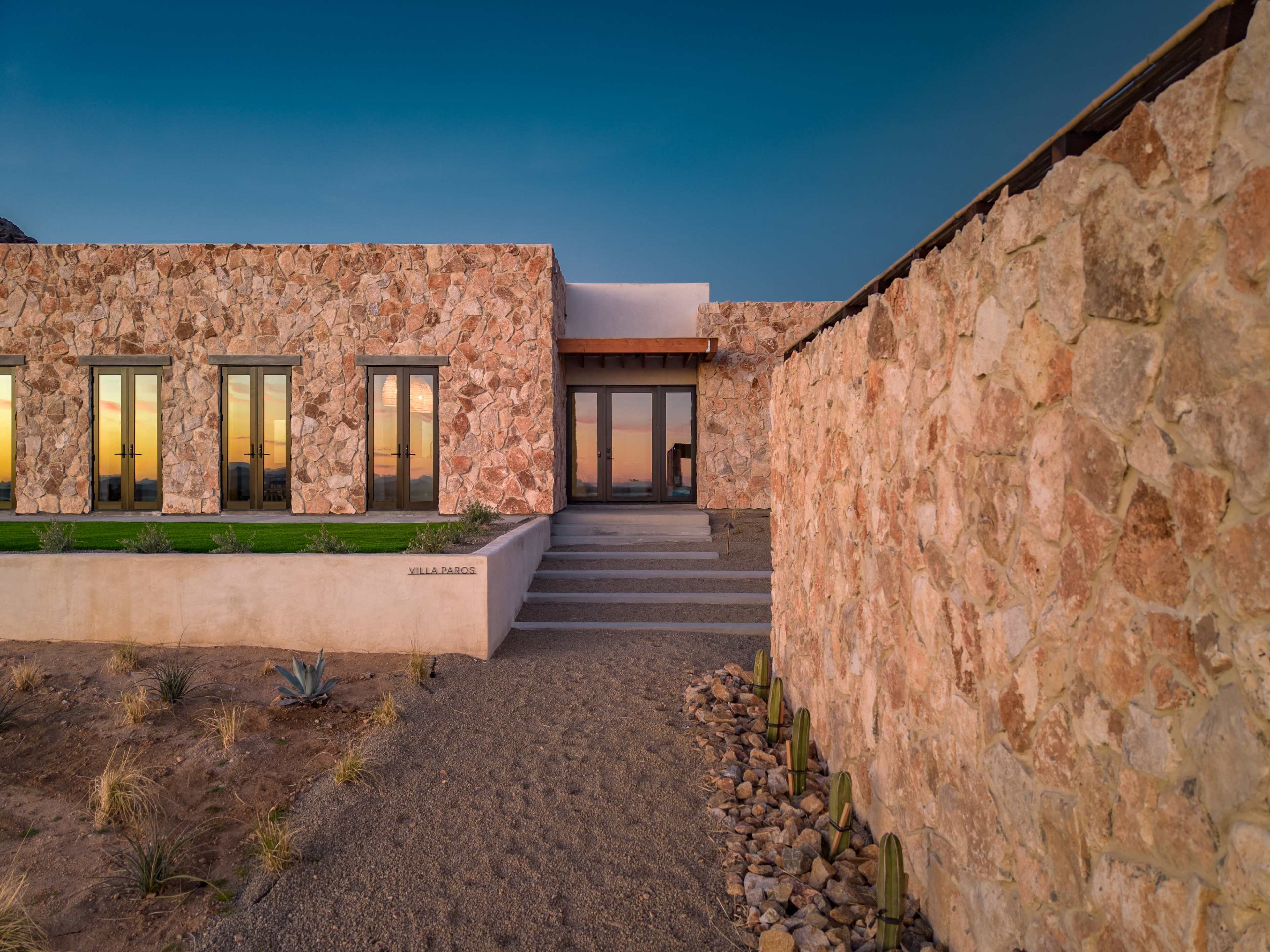 A modern stone-clad house with large glass windows is set against a twilight sky, featuring a neatly landscaped entrance.