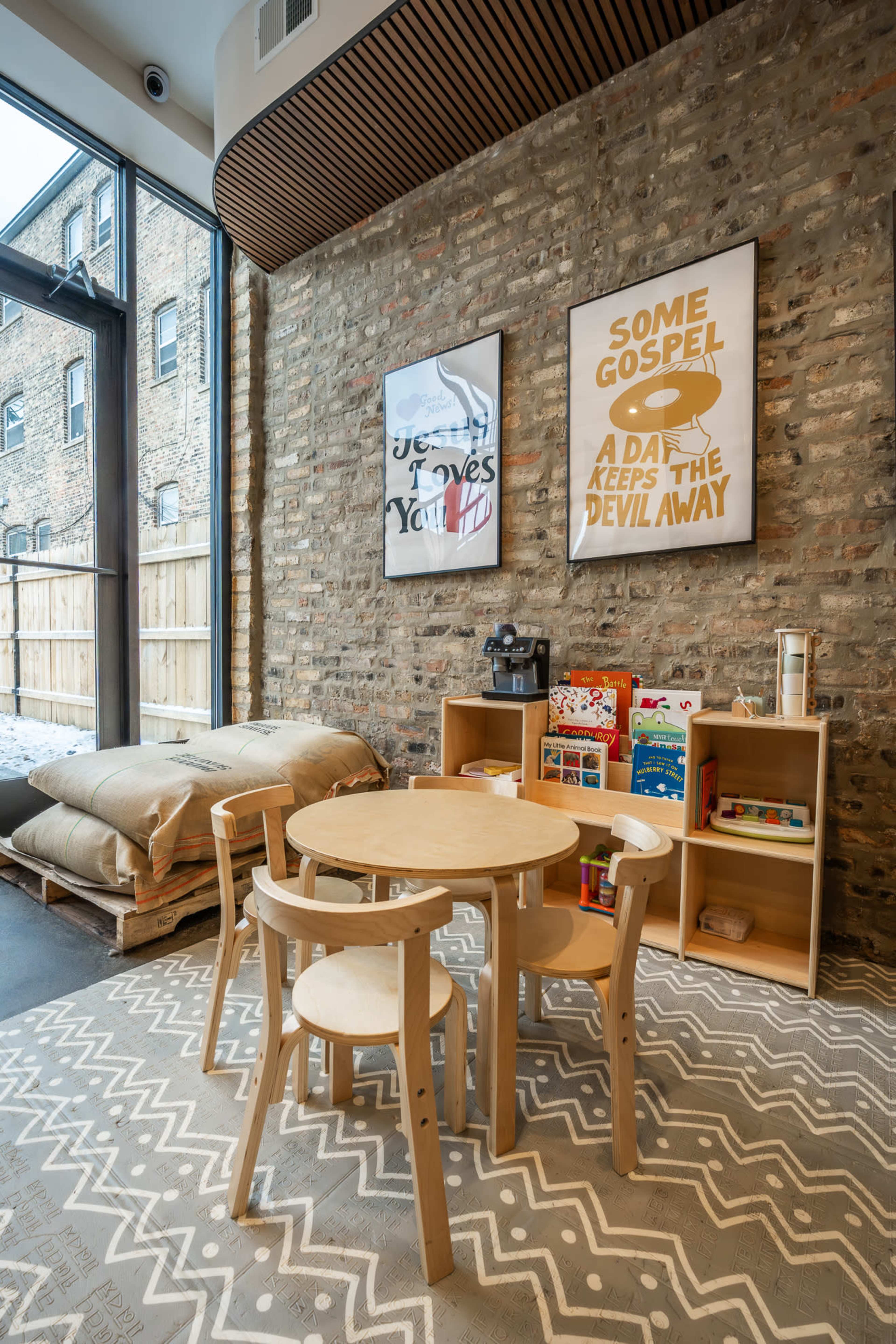 The image shows a cozy room with a stone wall, featuring a wooden table and chairs, a shelving unit filled with books and toys, and two framed posters on the wall.