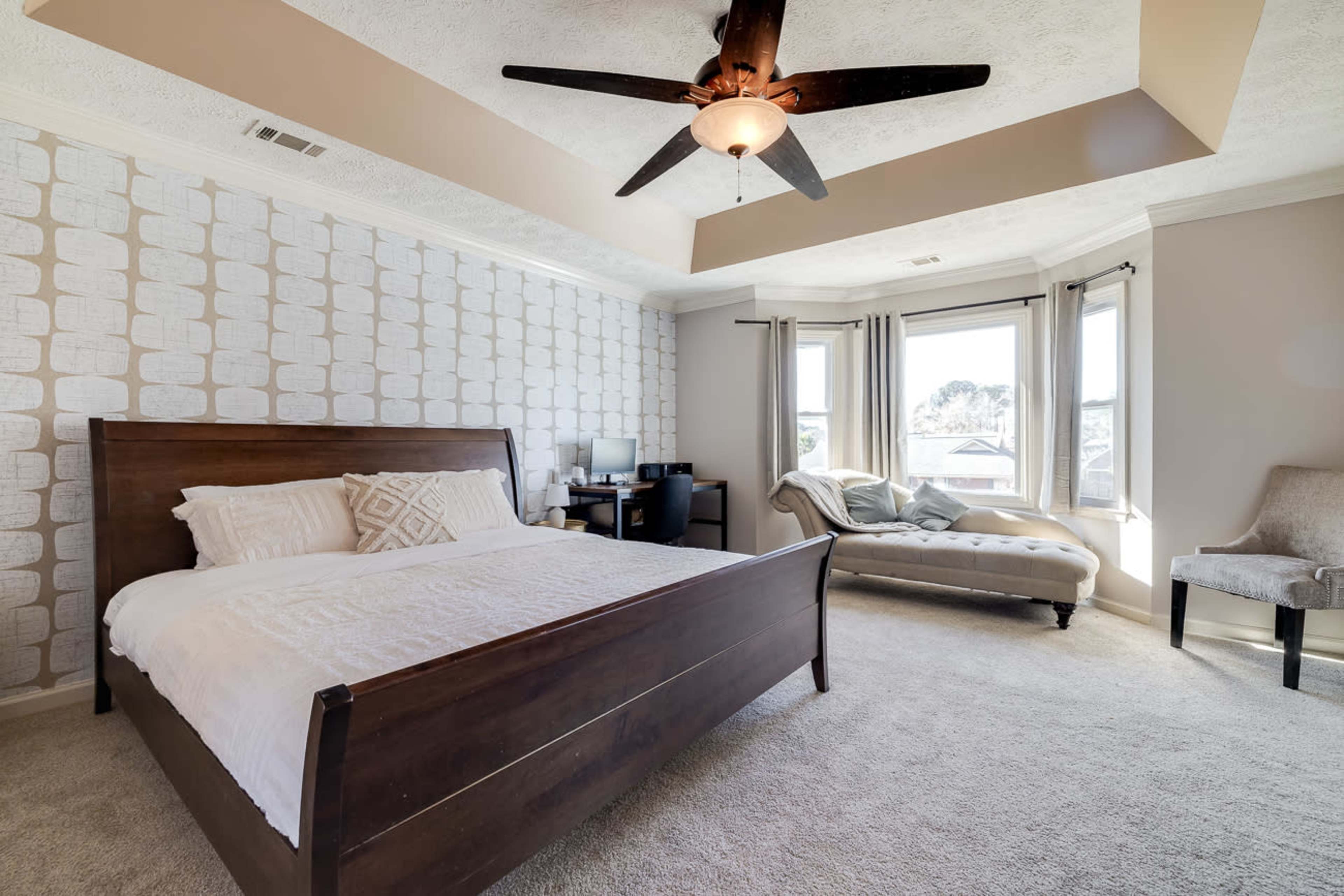 A bedroom featuring a wooden bed, a cozy seating area, a desk with a computer, and large windows allowing natural light to enter.