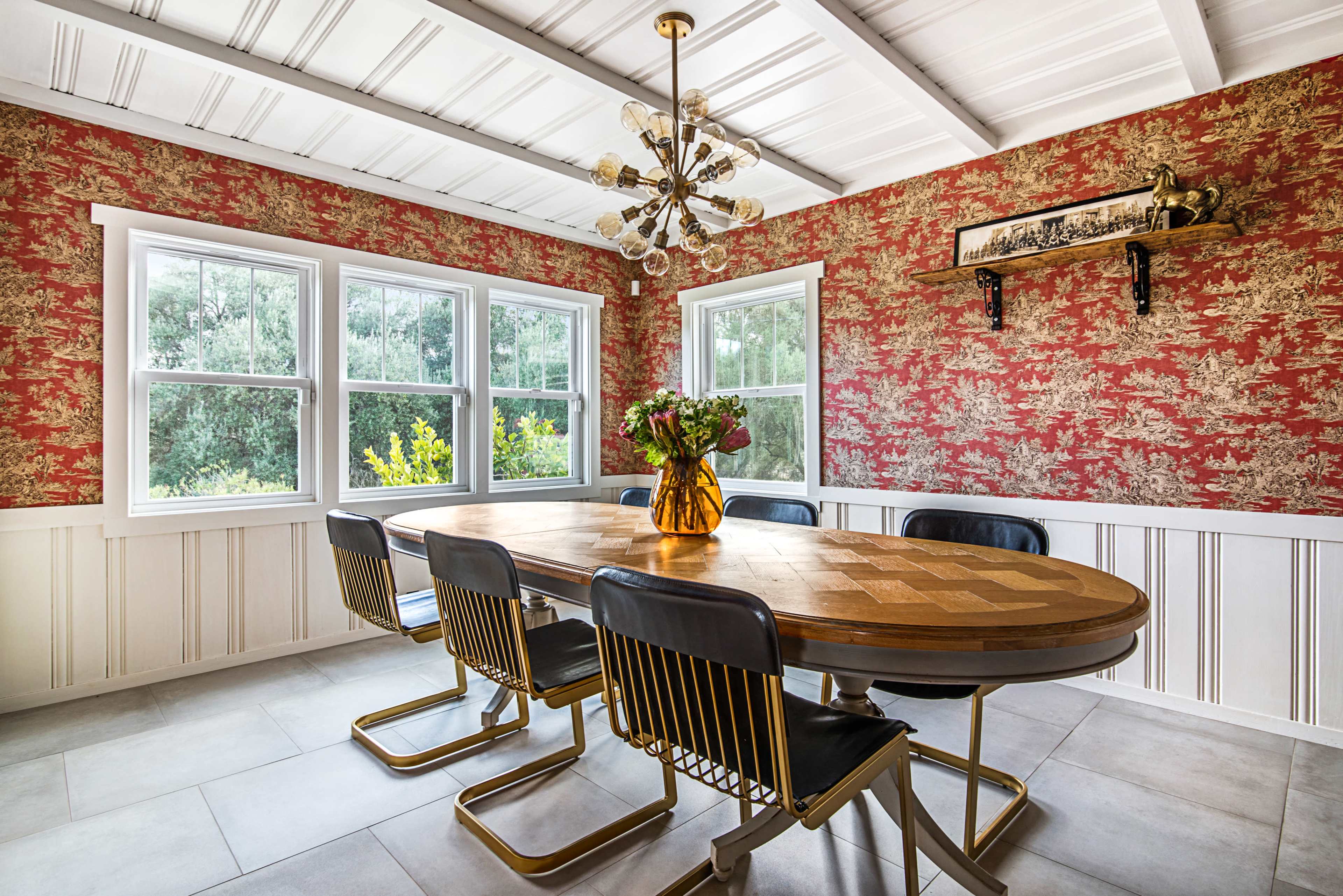 A dining room features a wooden table surrounded by black chairs, with a floral-patterned red wallpaper and large windows letting in natural light.