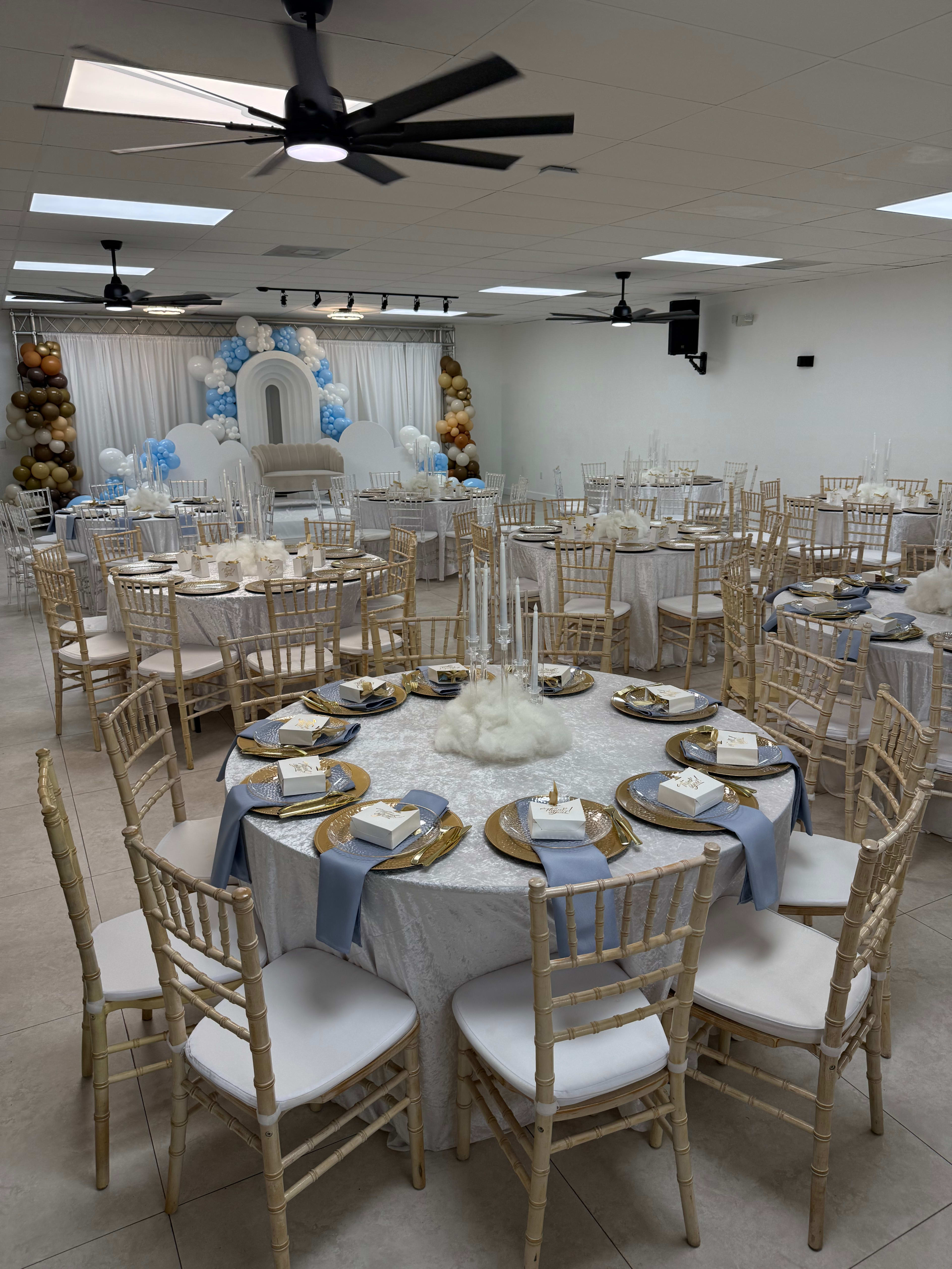 The image shows a decorated event space with round tables set for a formal gathering, featuring white tablecloths, gold and blue accents, and floral arrangements.