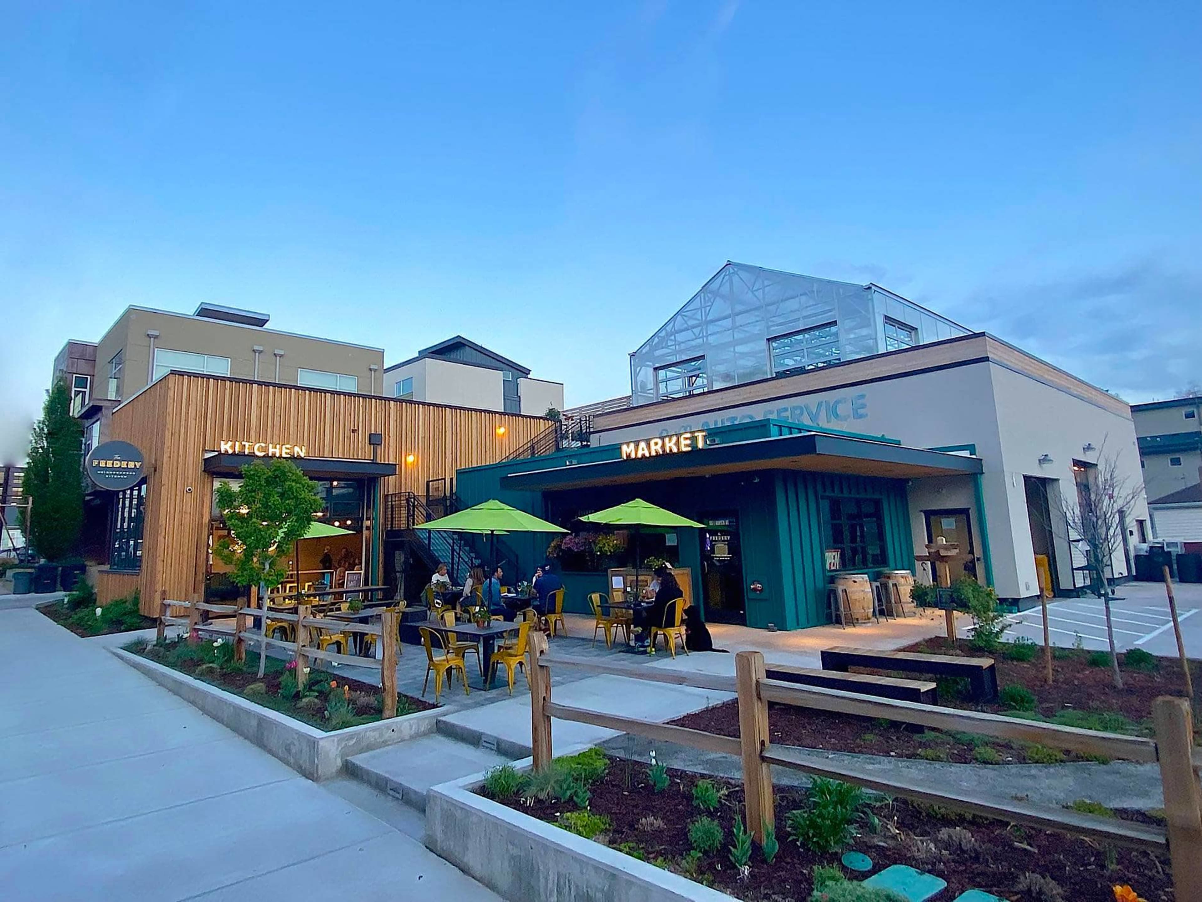 The image shows a modern building complex featuring a kitchen and market area with outdoor seating and green umbrellas.