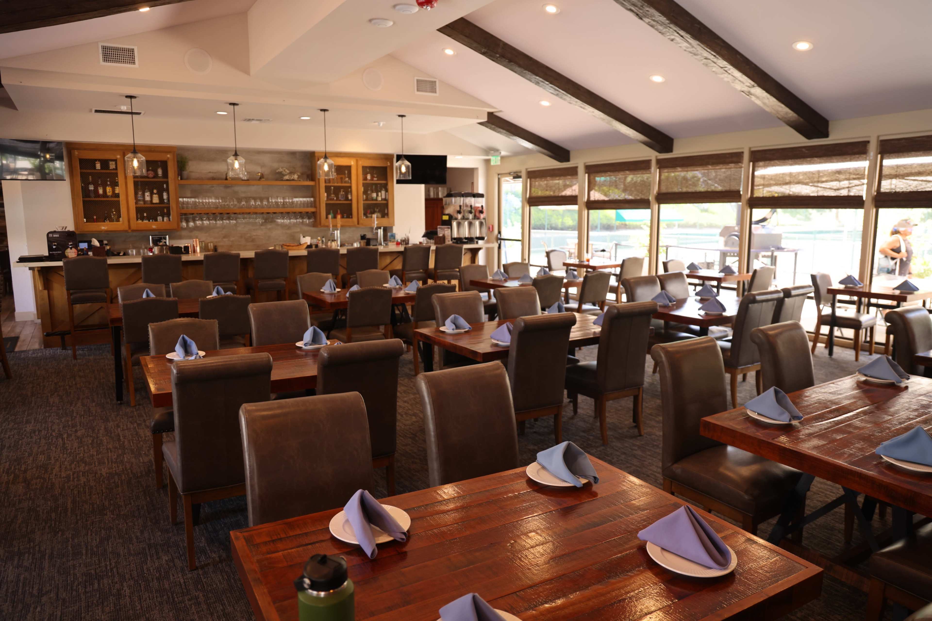 The image shows a restaurant interior featuring neatly arranged tables with blue napkins and a bar area in the background.