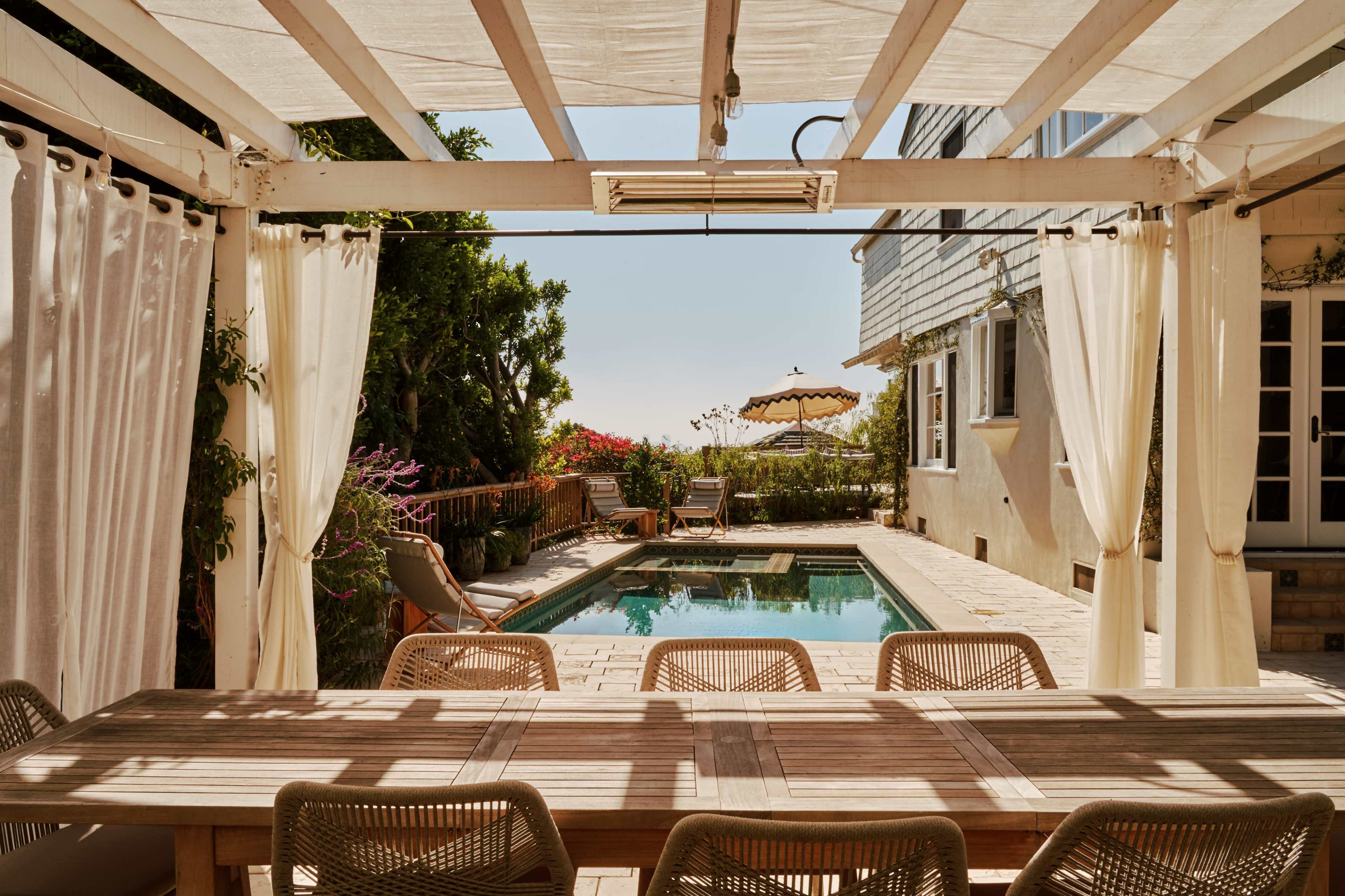 A wooden dining table with woven chairs is set under a pergola next to a pool, with sun loungers and potted plants in the background.