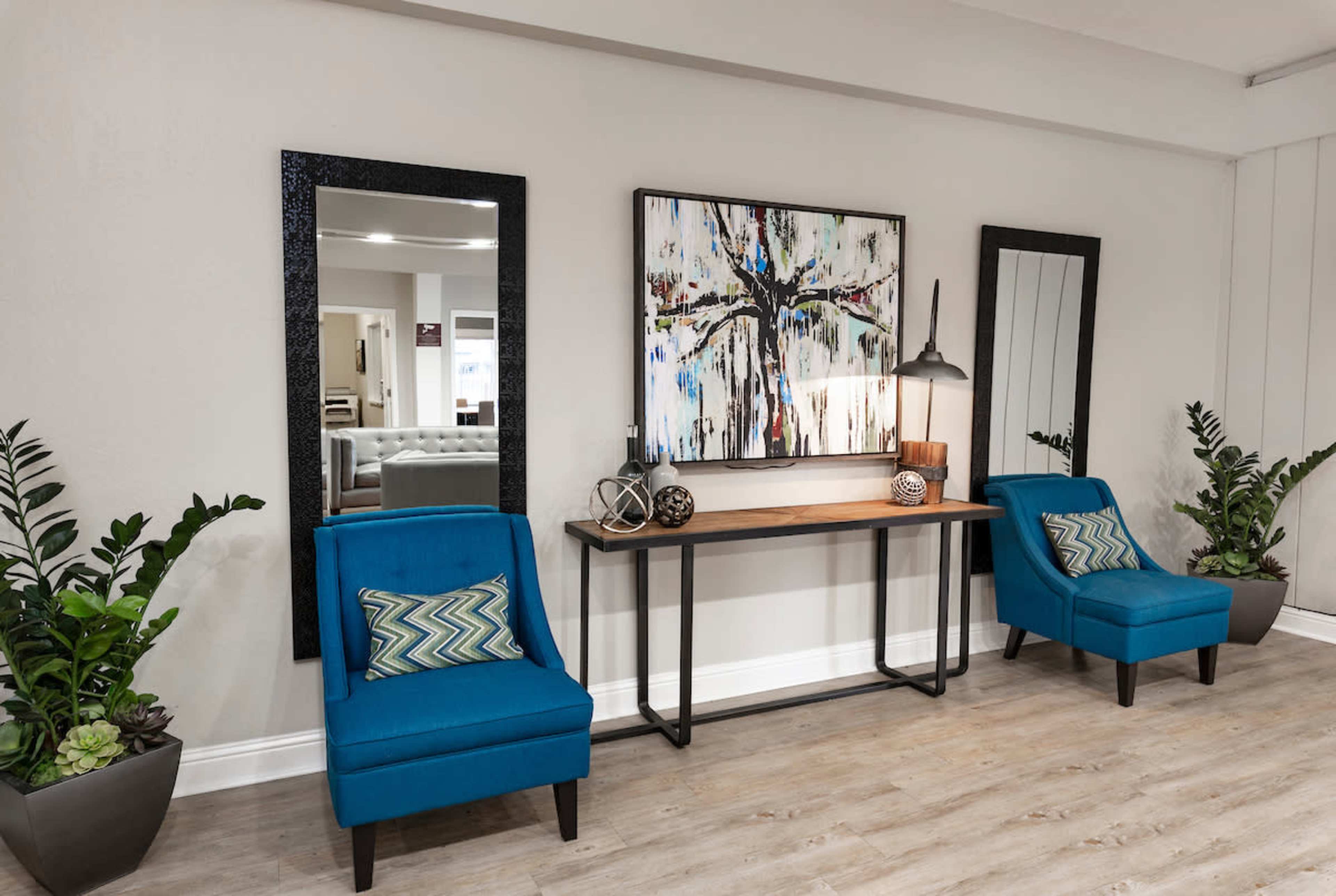 A modern entryway with two blue chairs, a wooden console table, and decorative plants flanking the sides, alongside two mirrors on the wall and artwork above the table.