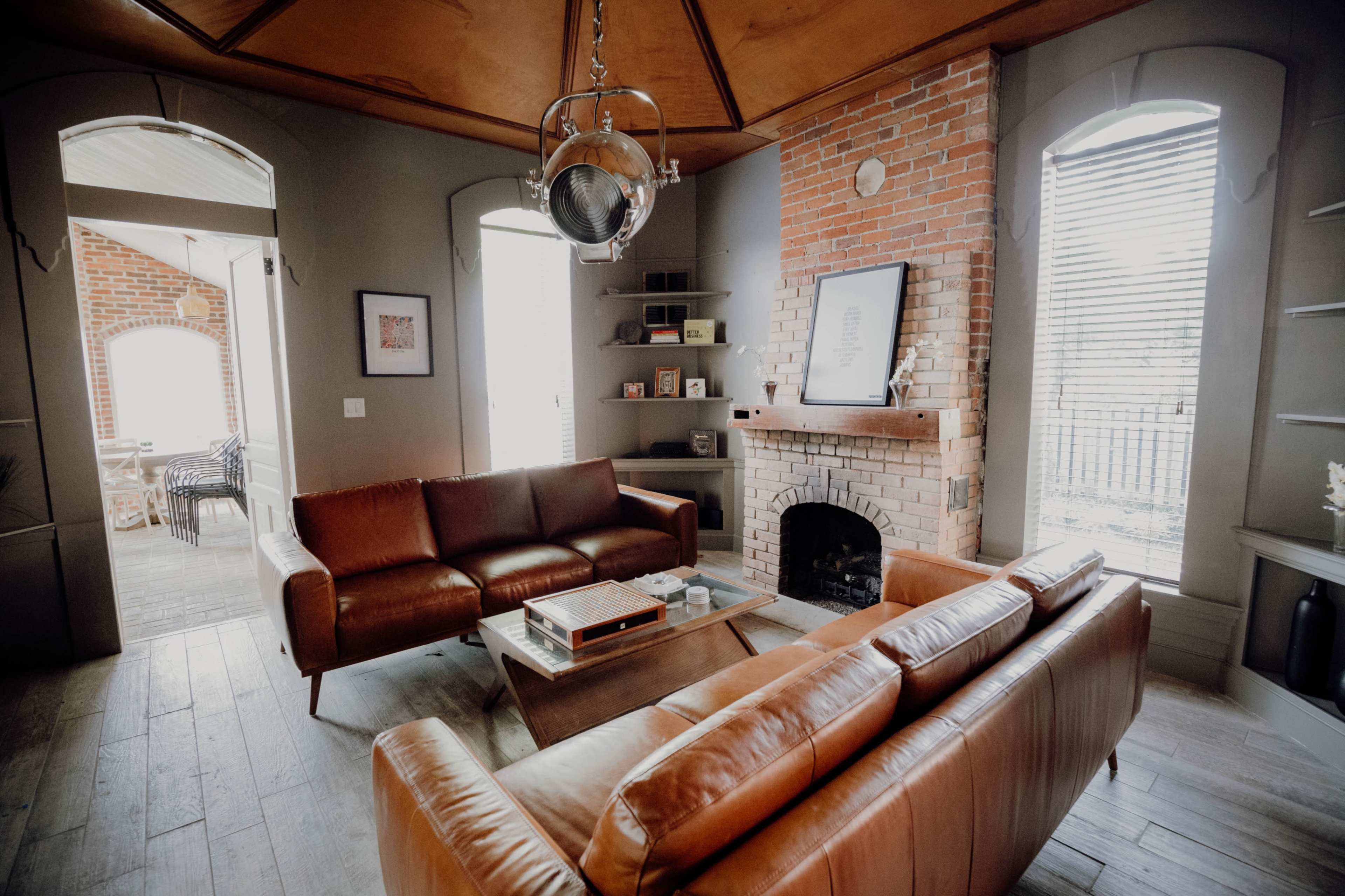 A modern living room with brown leather sofas, a central coffee table, a brick fireplace, and large windows allowing natural light to enter the space.