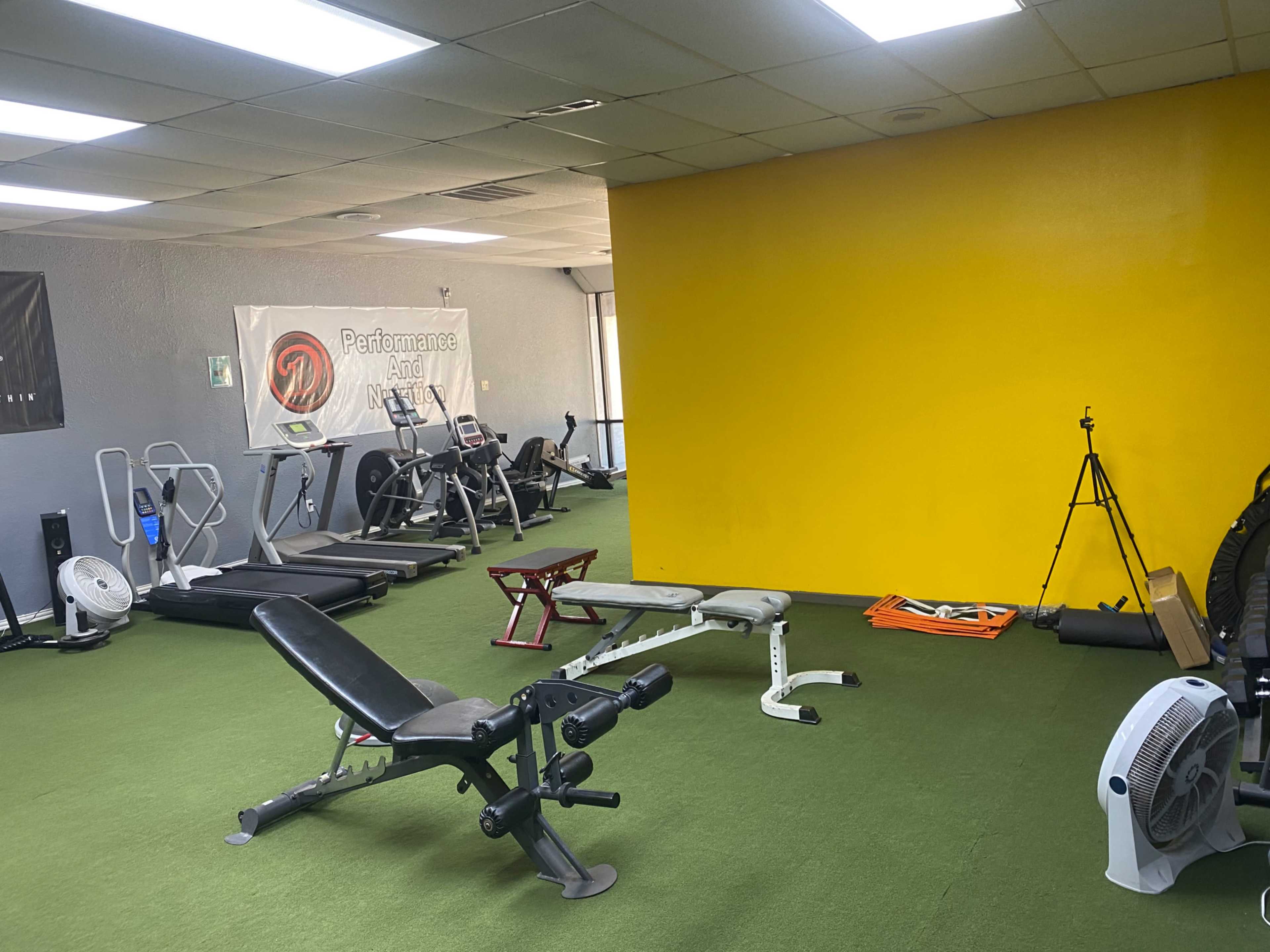 A gym interior with various exercise equipment, green flooring, and a yellow wall in the background.