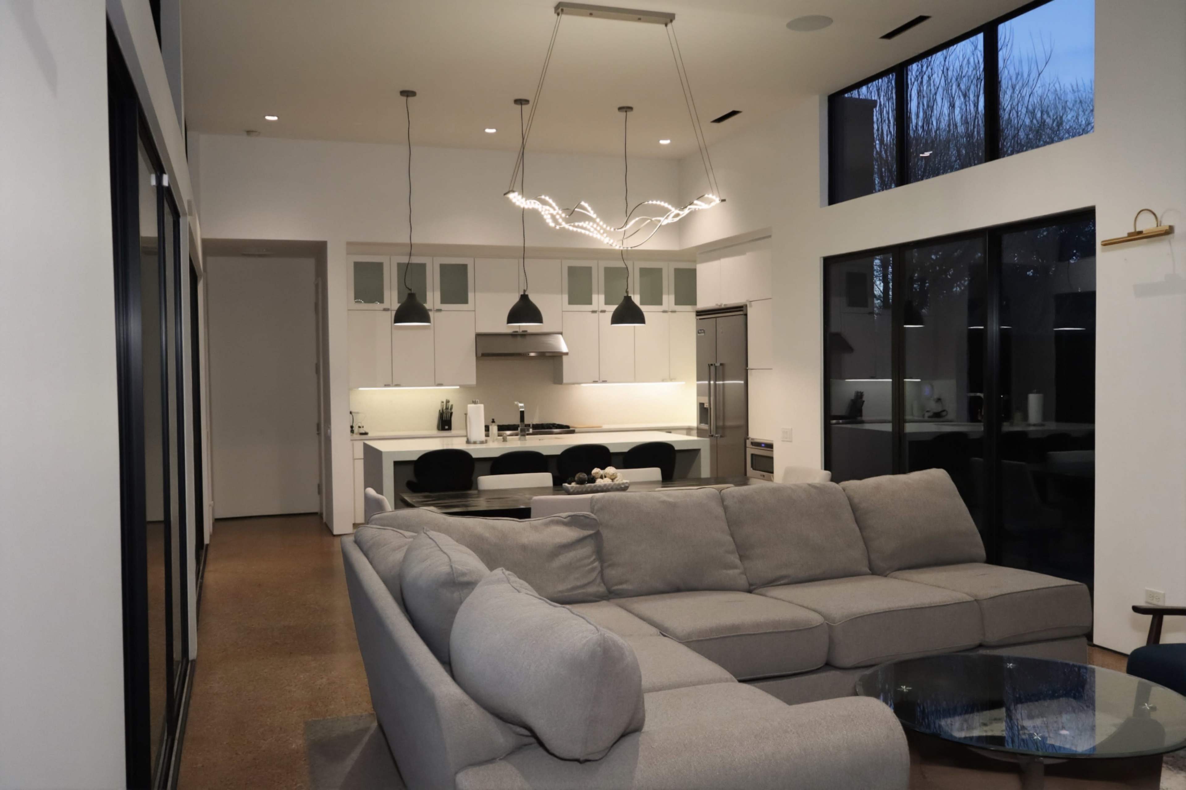 The image shows a modern living room featuring a large gray sectional sofa, a glass coffee table, and an open kitchen area with pendant lighting and bar seating.
