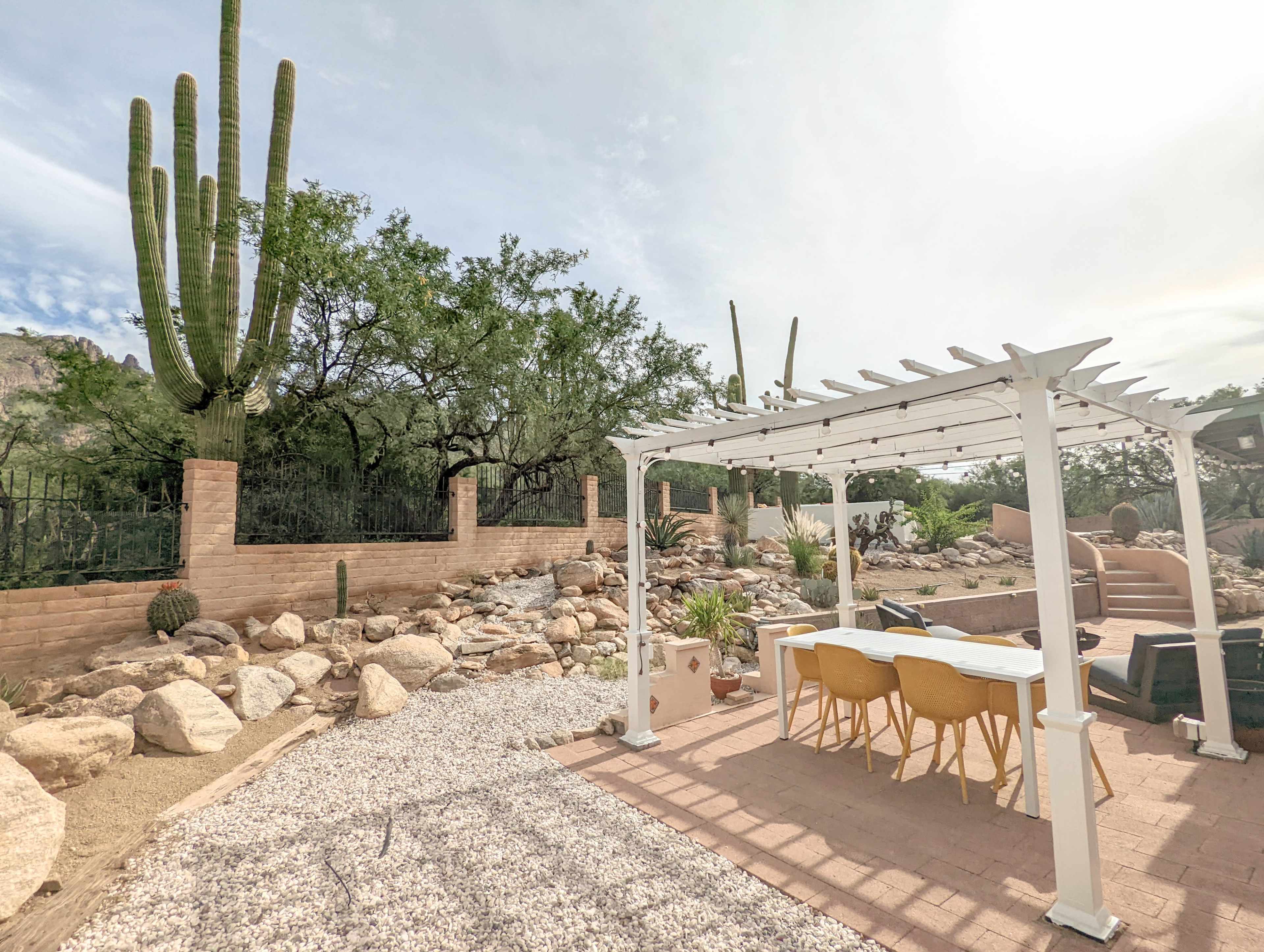 A pergola with a dining table and chairs is set on a stone patio surrounded by desert plants and cacti.