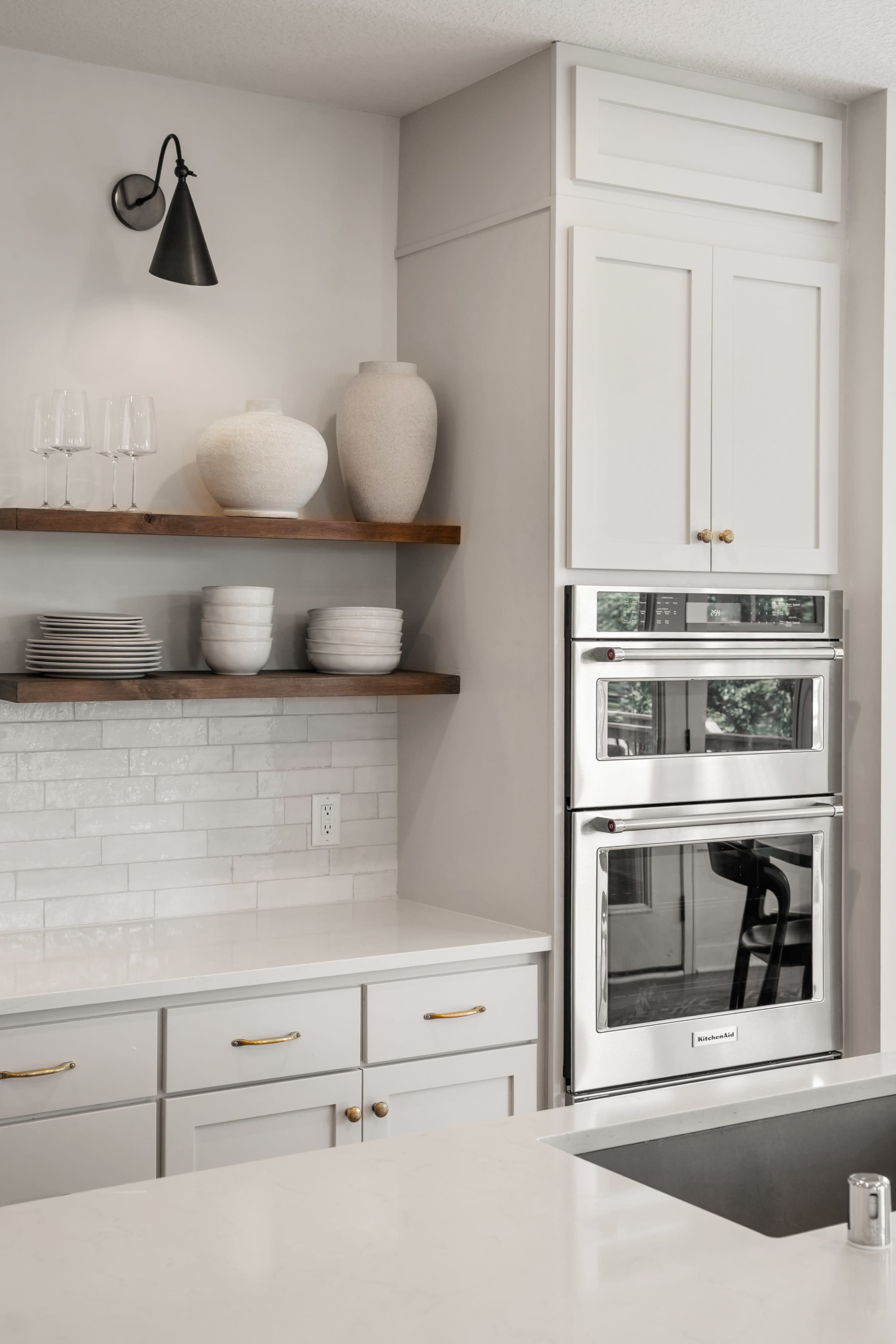 The image shows a modern kitchen featuring white cabinetry, open shelving with dishware, and stainless steel appliances stacked vertically.