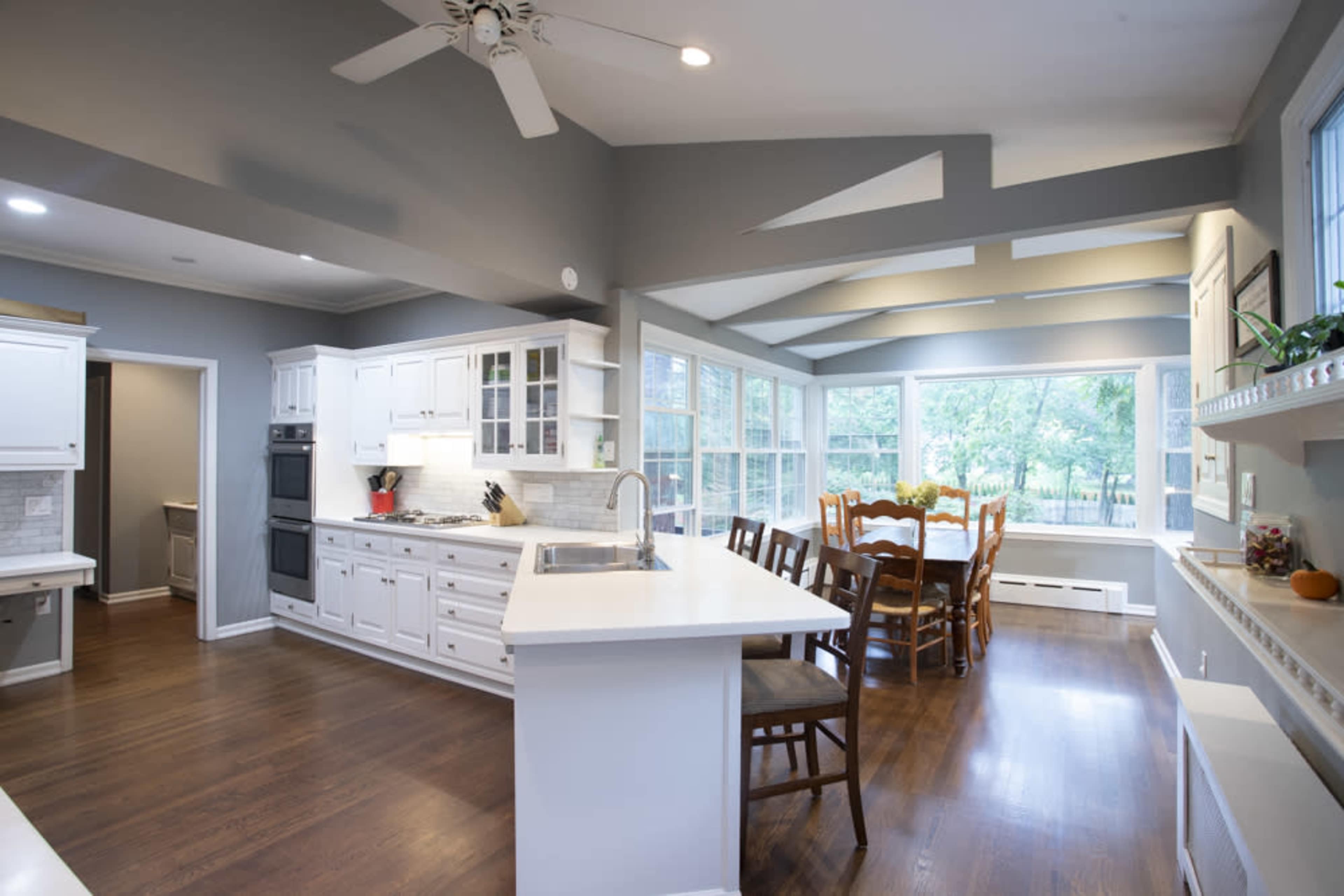A modern kitchen features white cabinetry, a large island with a sink, and a dining area illuminated by natural light from large windows.