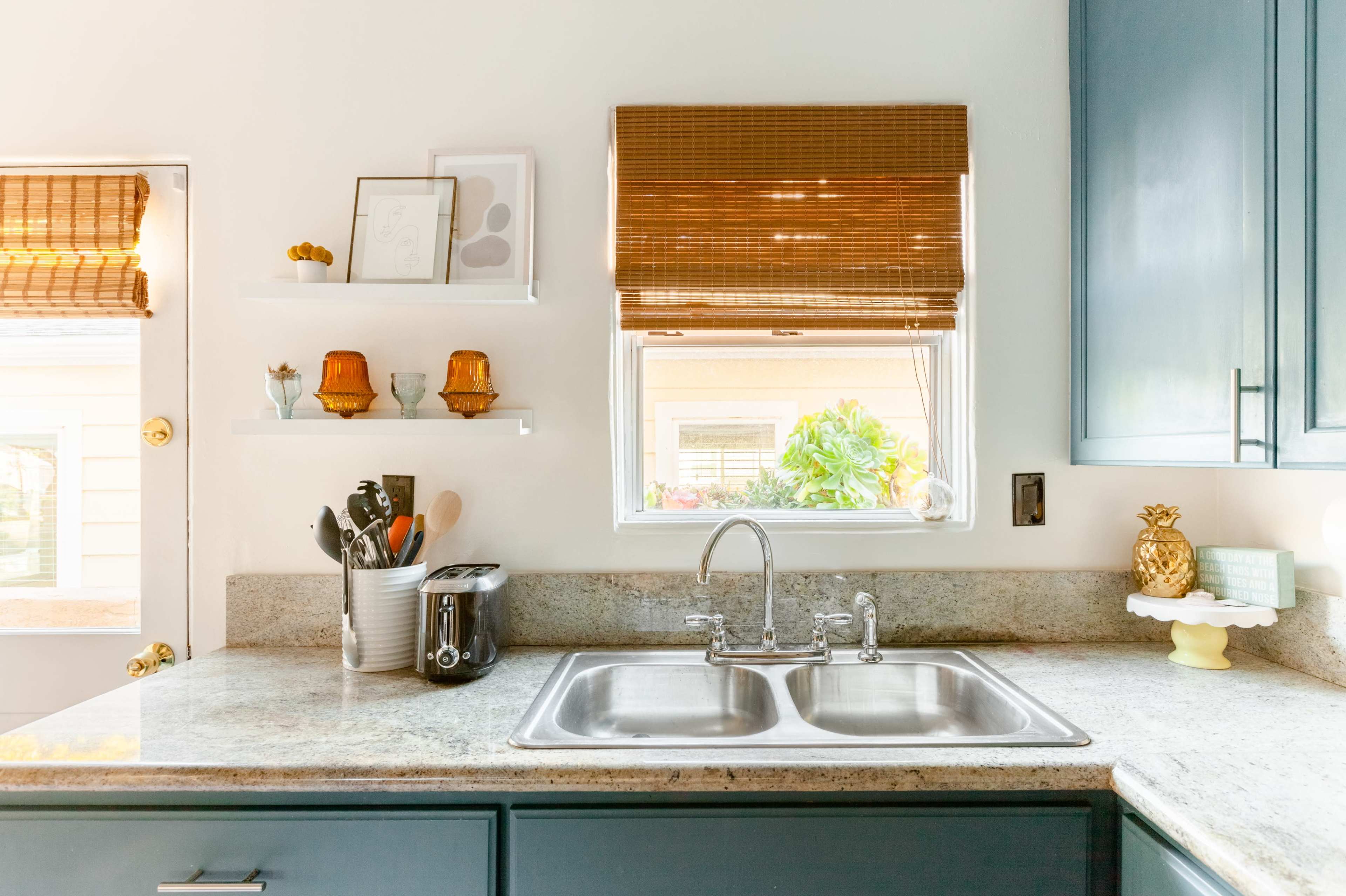 A kitchen features a double sink, granite countertop, and a window with bamboo shades, along with various utensils and small decor items on the shelves.