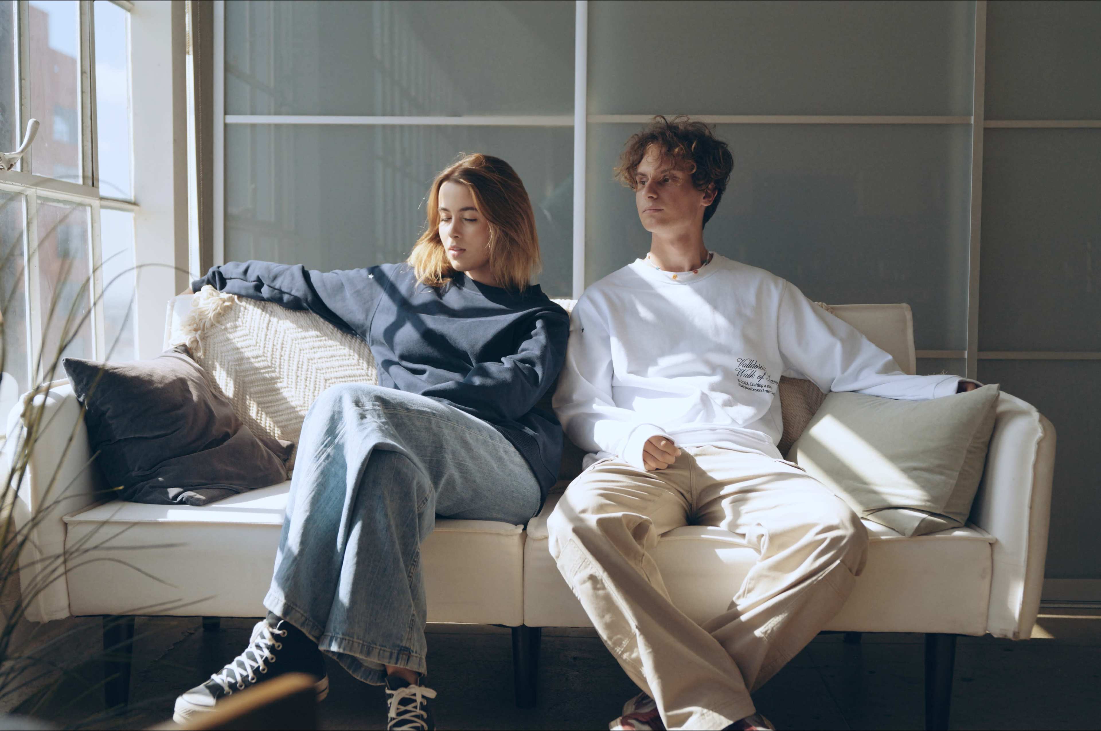 Two young adults sit apart on a couch in a sunlit room, wearing casual clothing.