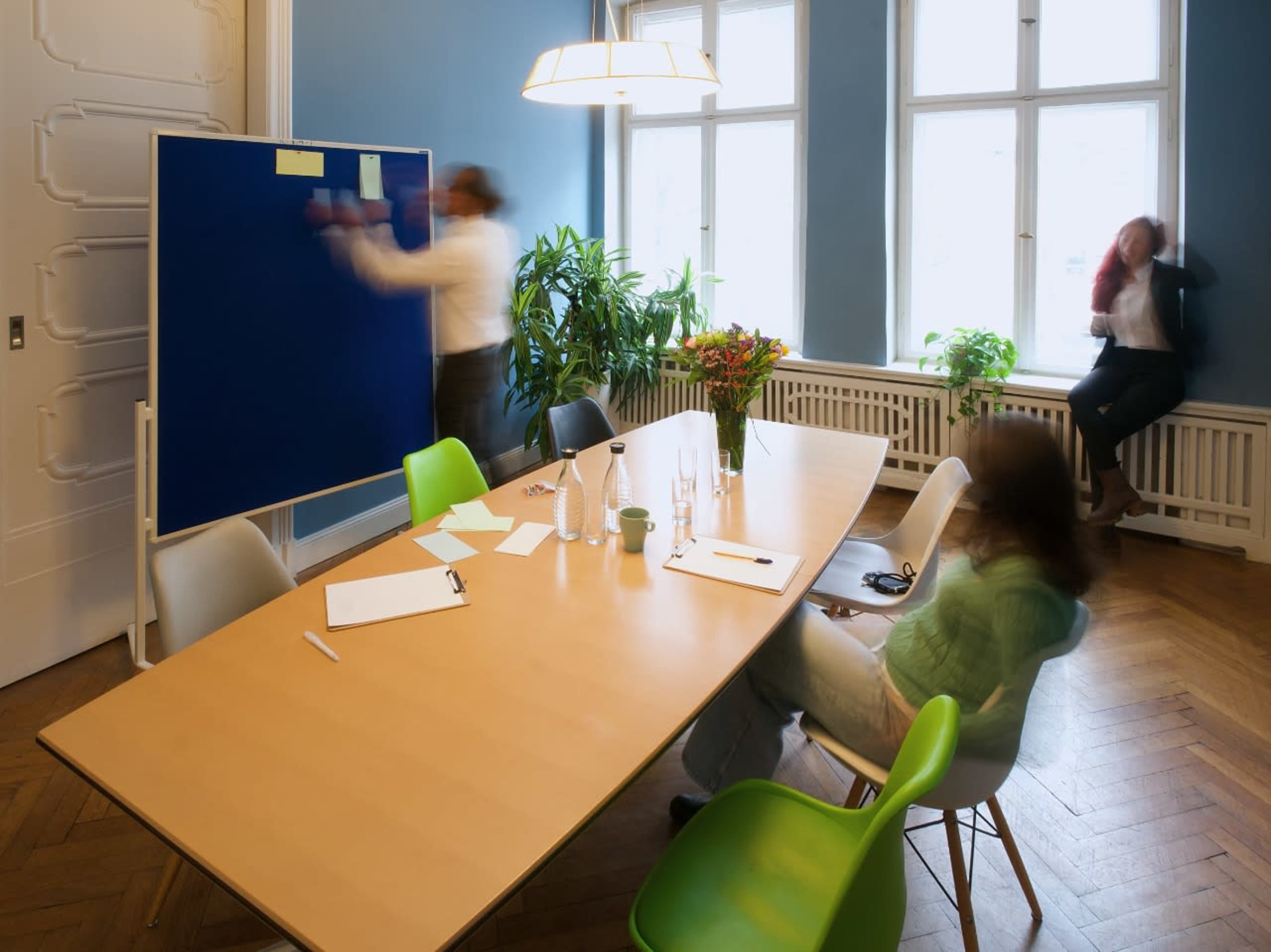 A group of people is engaged in a meeting around a wooden table in a brightly lit room, with one person writing on a blue board while another is seated and observing.