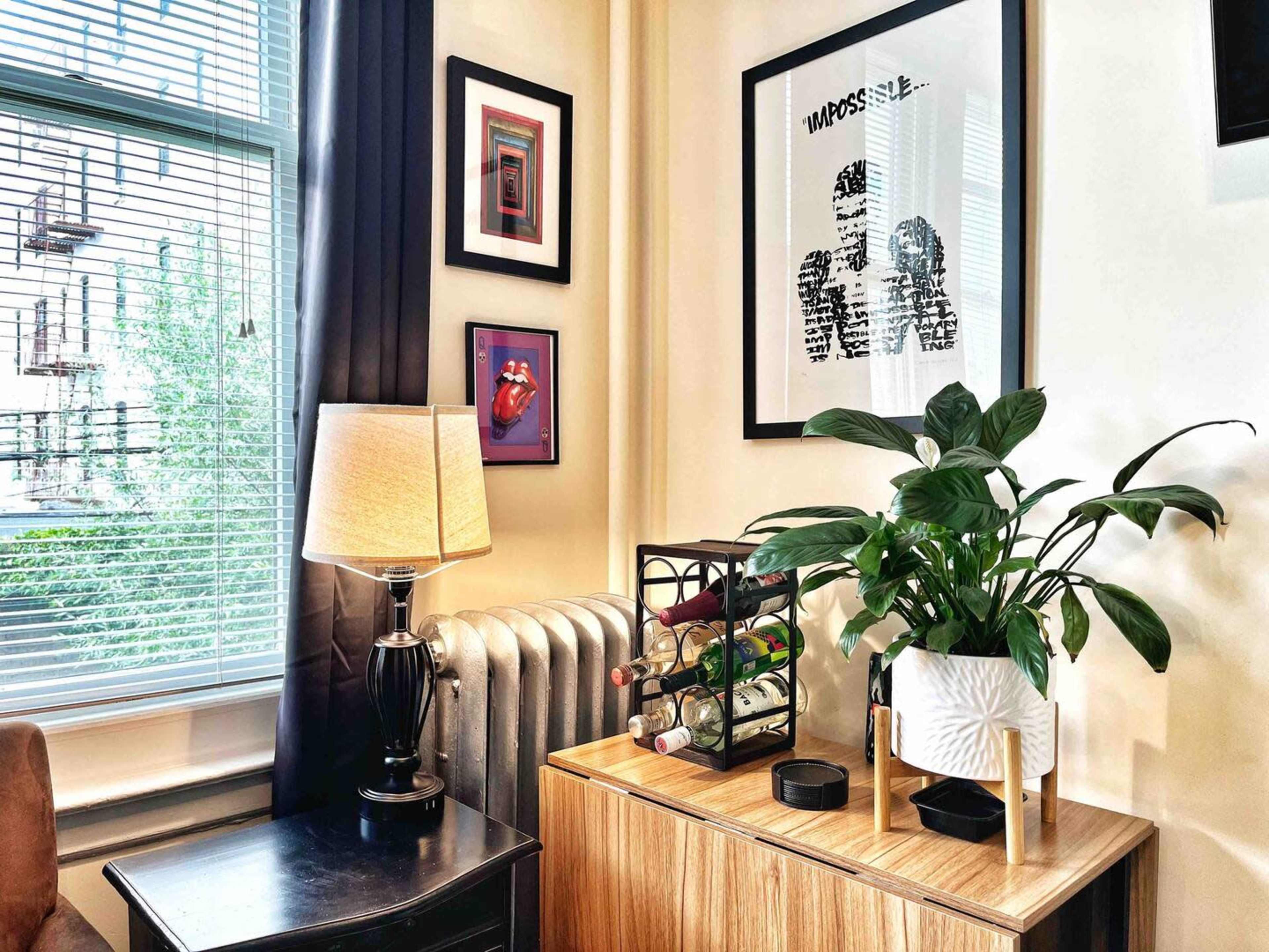 A cozy living room corner with a lamp, framed art on the wall, a plant, and a wine rack atop a wooden cabinet.