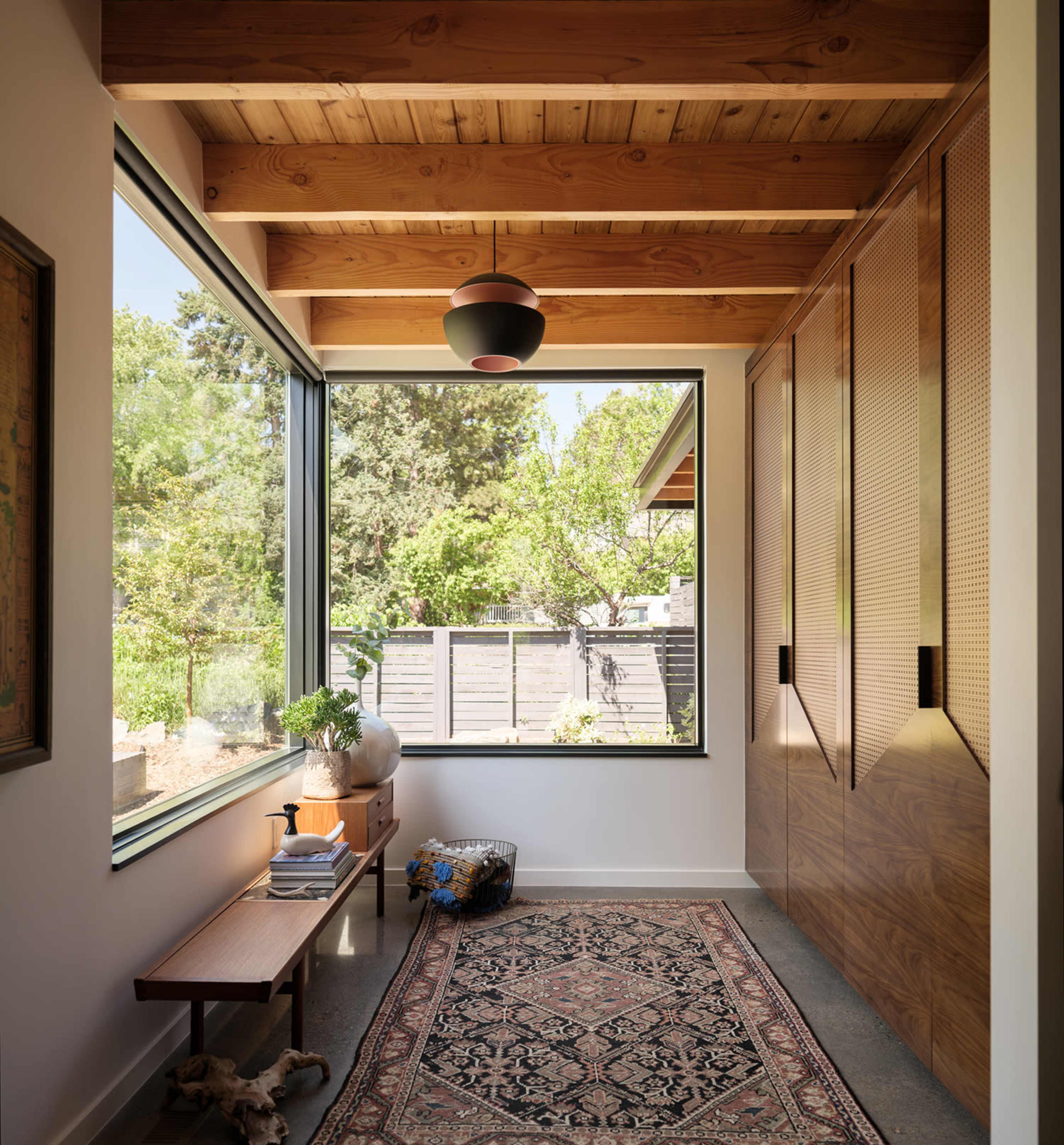 The image shows a modern entryway featuring large windows, wooden ceiling beams, a narrow bench, and an area rug on a concrete floor.