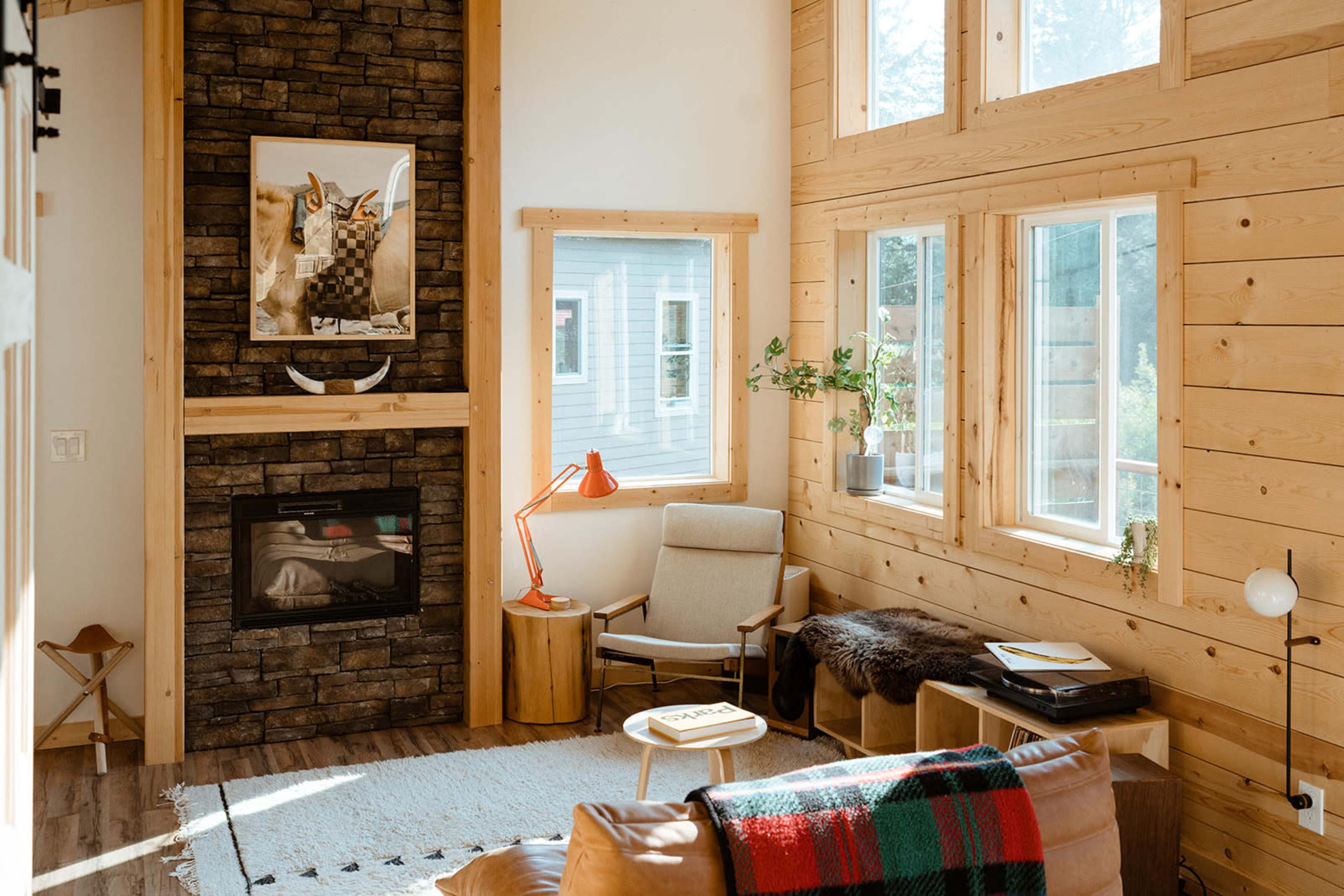 The image shows a cozy living room with wood paneling, a stone fireplace, large windows allowing natural light, and minimalistic furnishings including a chair, a side table, and a record player.