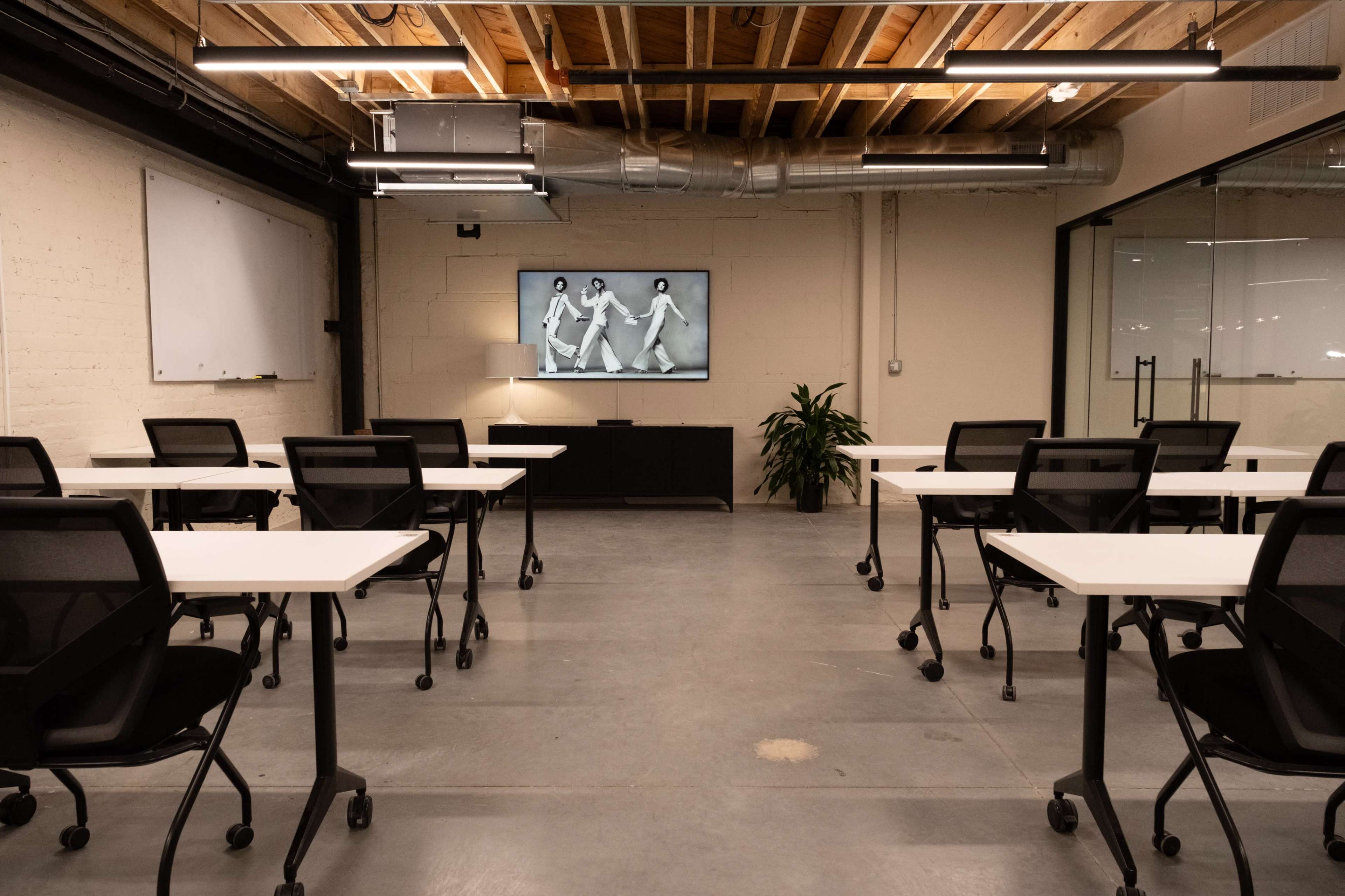 The image shows a modern classroom with several movable chairs and tables arranged around the room, featuring a large screen displaying a black-and-white image on one wall.