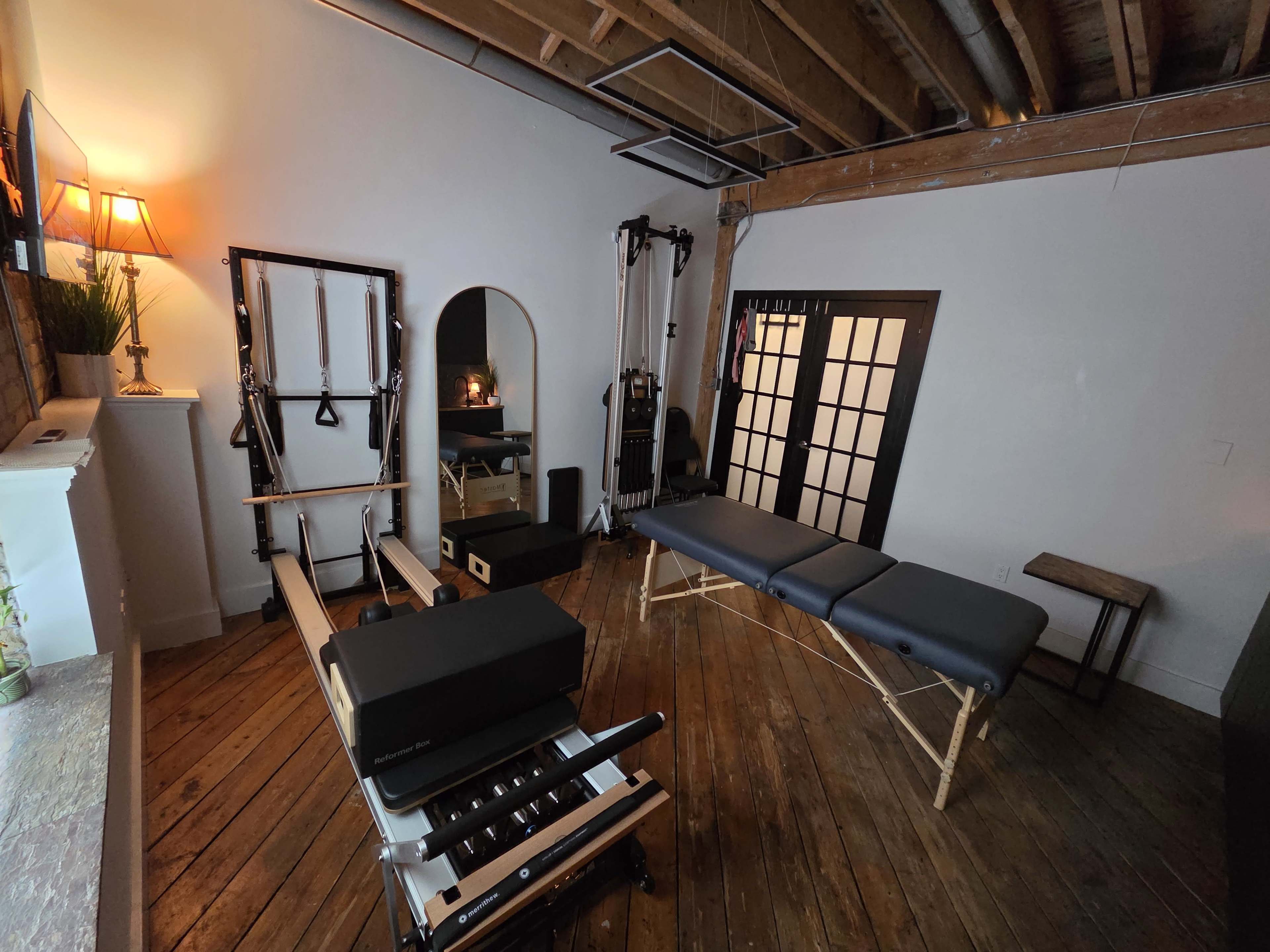 The image shows a spacious room featuring a Pilates reformer, a treatment table, and various fitness equipment, all situated on wooden flooring.