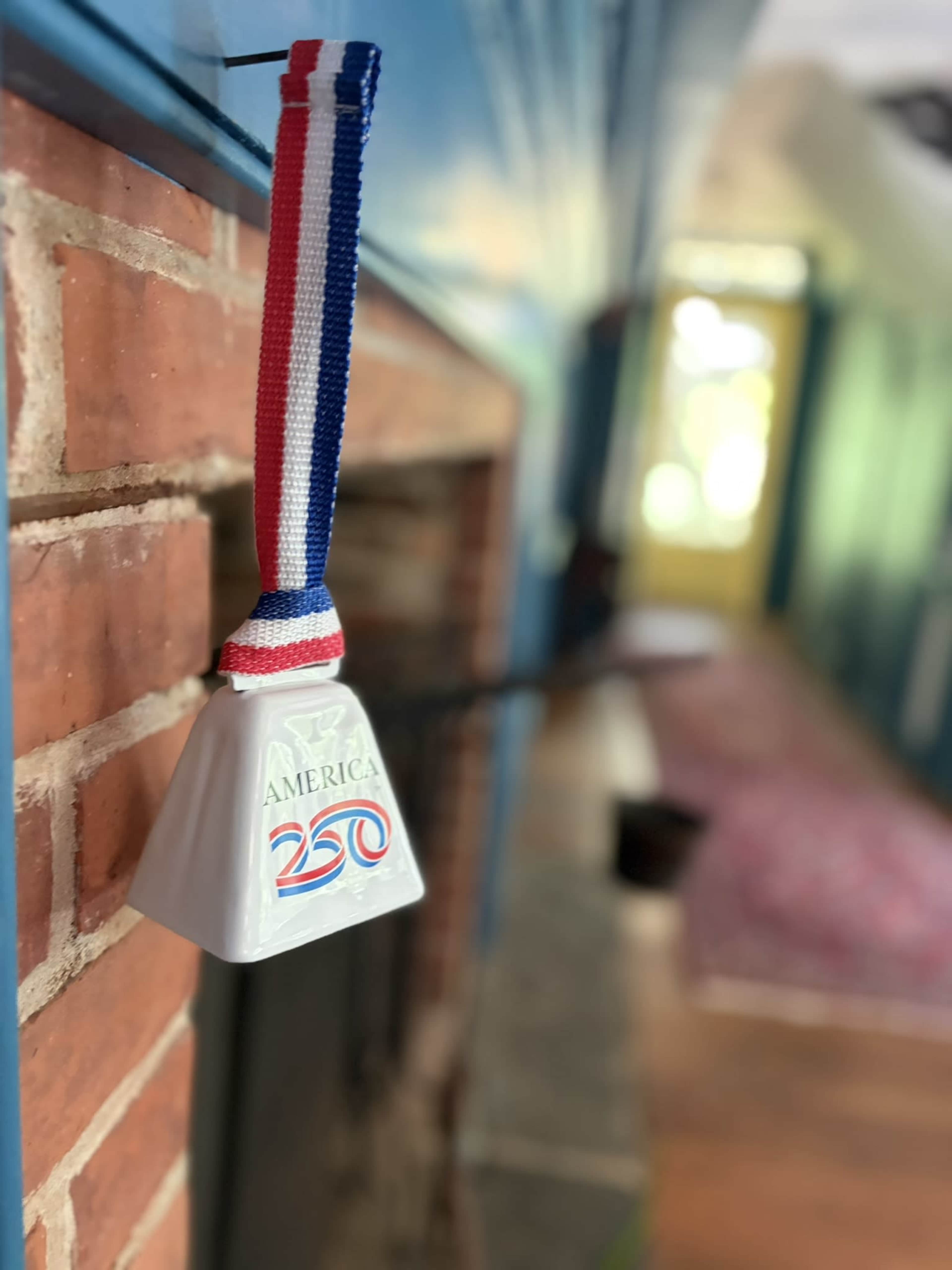 A ceramic bell with a red, white, and blue ribbon hangs from a brick wall in a brightly lit hallway.
